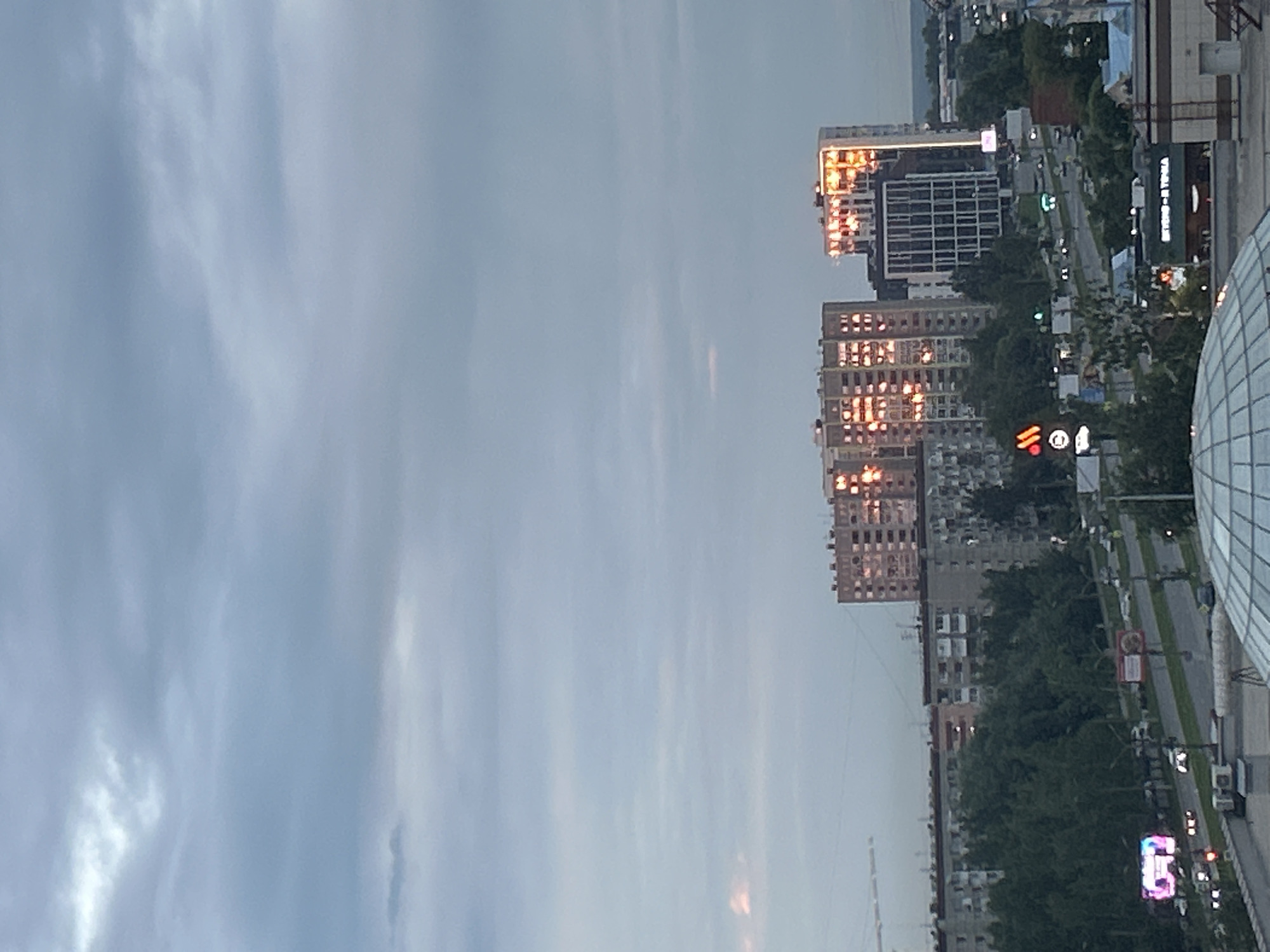 The image depicts an urban skyline in Izhevsk, Russia, under a cloudy sky with sunlight reflecting off buildings. In the mid-ground, two multi-story apartment buildings are prominent, their windows illuminated by the setting sun, creating a warm, orange glow. To the left of these buildings are more apartment blocks and a dense line of trees. In the foreground, a glass dome structure is partially visible, suggesting a viewpoint from a building or public space. A road runs through the scene, with several cars and a few streetlights visible. Several signs are present, including a red and orange circular logo that resembles the McDonald's logo, and another sign with Cyrillic text that reads "BERRY - TAKA". The overall scene captures a moment of urban life as day transitions to evening.