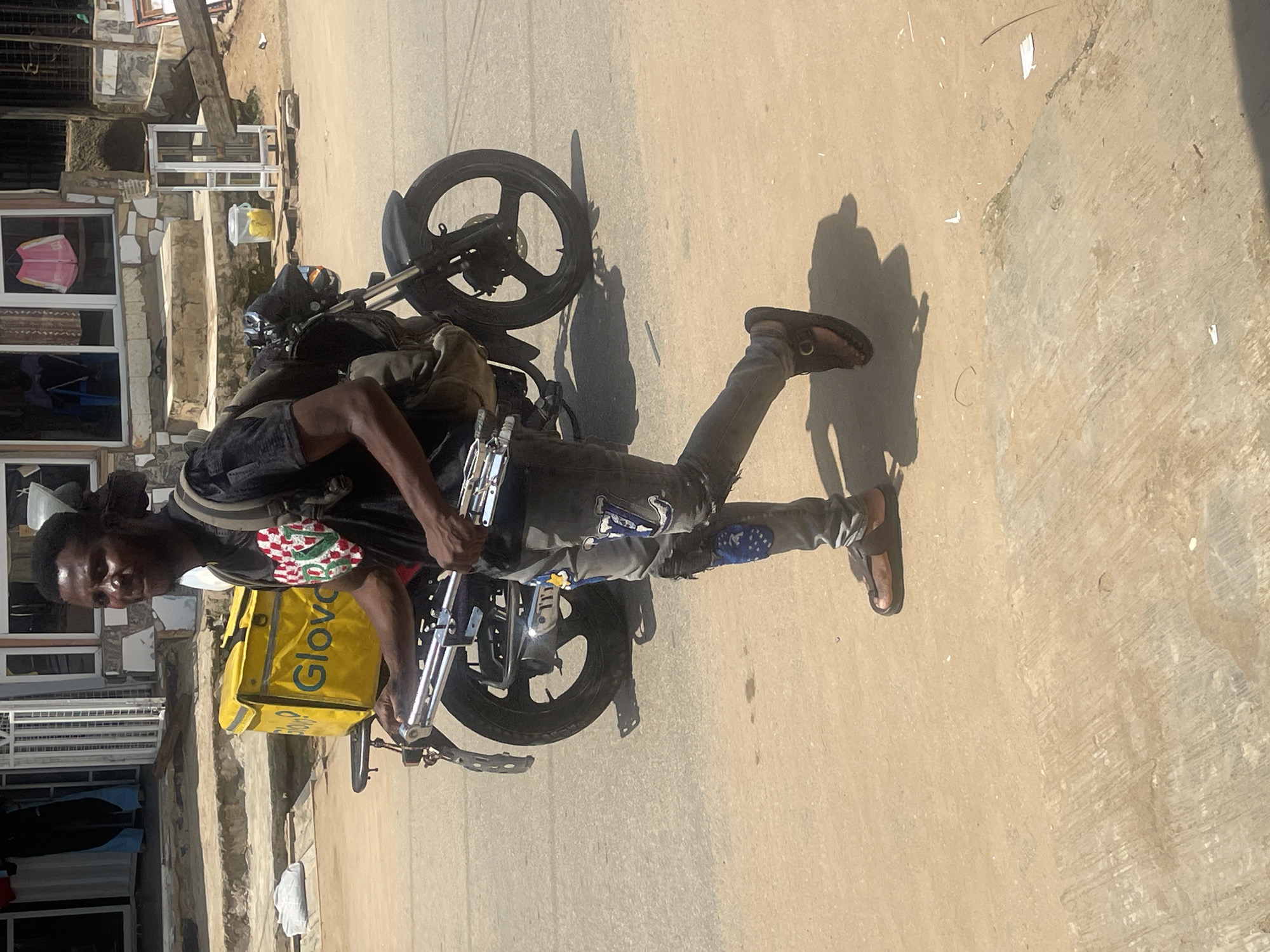 A young man is standing next to a black motorcycle in what appears to be a residential area in Abuja, Nigeria. He is wearing a black t-shirt with a red and white checkered patch on the left breast and dark pants with blue and white patterned patches. He has on brown sandals. The man has a yellow delivery bag on the back of the motorcycle, which has the word "Glovo" printed in dark blue letters. The bag is partially obscured by the man.
The scene appears to be outdoors, with sunlight casting shadows on the paved ground, indicating it is daytime. The background shows a building with several windows, some of which are boarded up or have grates. Steps lead up to the building. There is some debris on the ground. The man is looking towards the camera with a neutral expression. The overall impression is of a delivery person taking a moment pause.