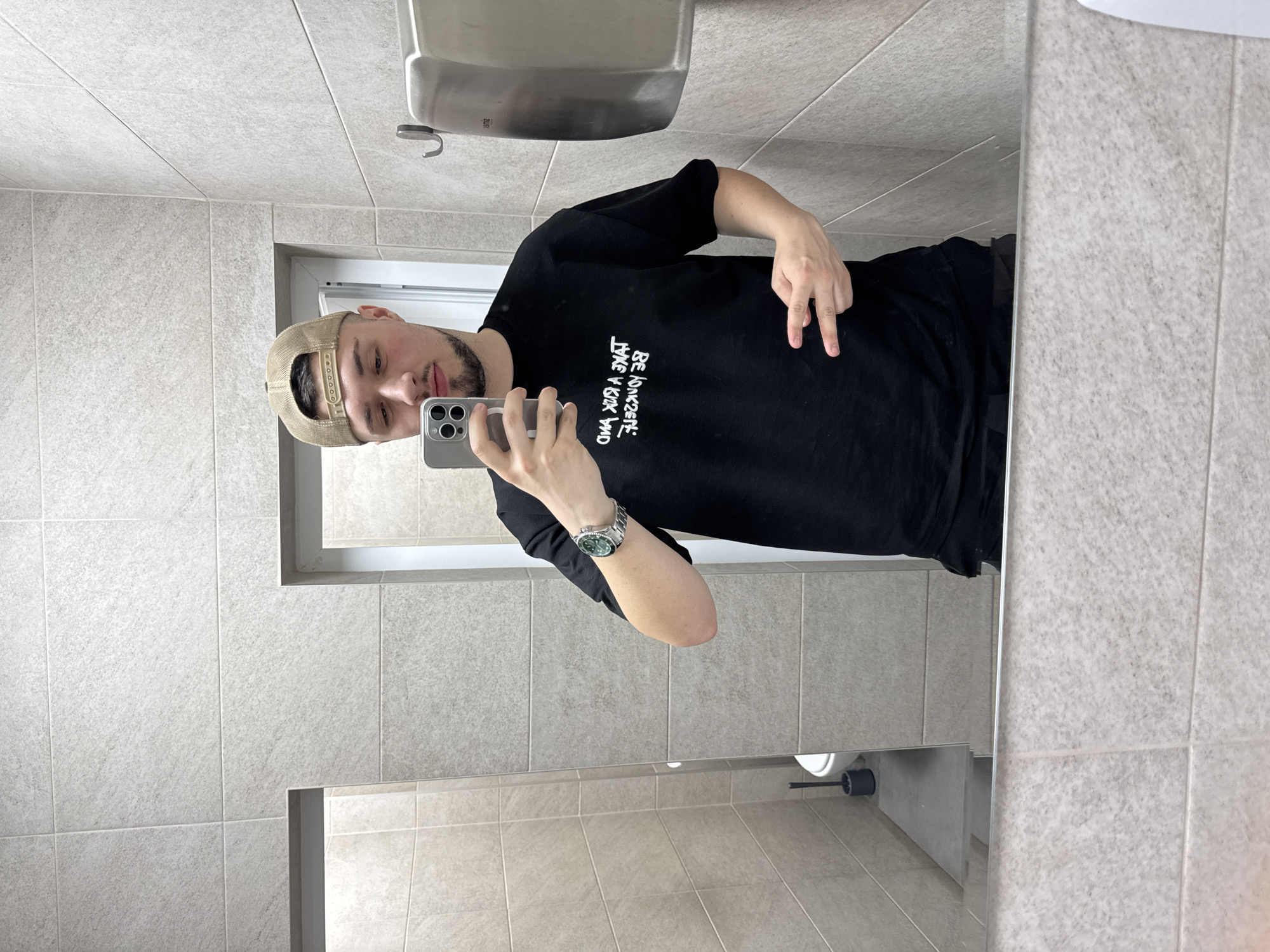 A man is taking a selfie in a bathroom. He is wearing a black t-shirt, black pants, and a beige baseball cap. He is holding a smartphone in his left hand. He is making a hand gesture with his right hand. The bathroom has light gray tiles and a stainless steel hand dryer. There is a toilet visible in the reflection of the mirror. The location is unknown and cannot be pinpointed to the city of Varna, Bulgaria based on the provided image.