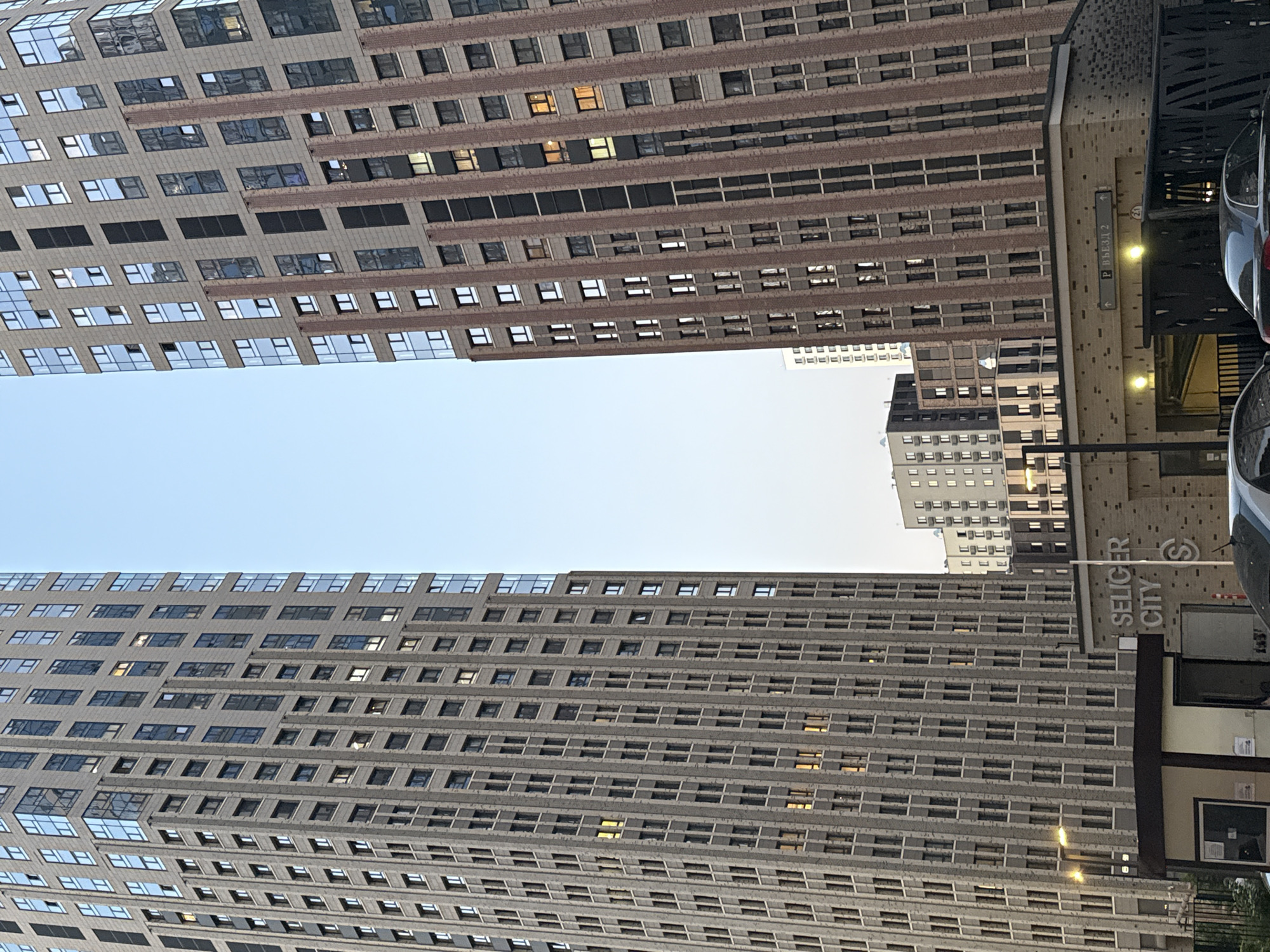 This photograph depicts two high-rise buildings in Moscow, Russia, likely residential or commercial structures, viewed from a low angle, making them appear as if they are converging toward the top of the frame, where there is a clear sky. The buildings are distinguished by their architectural design: one features a repetitive pattern of windows and horizontal lines of a light color with some warm colors visible inside, while the other has a similar window design but appears in a greyscale tone. The repetitive window design of the two towers and horizontal lines suggest the buildings may have been constructed in the 1960s. A sign, tilted to the right, is partially visible, with "SELIGER CITY" printed vertically. Adjacent to it, a building entrance marked as "P ВЪЕЗД 2" indicating the location of parking area entrance number 2, likely a parking entrance for the building, also suggests a presence of a parking garage. The cars can be seen in the right side of the frame.
