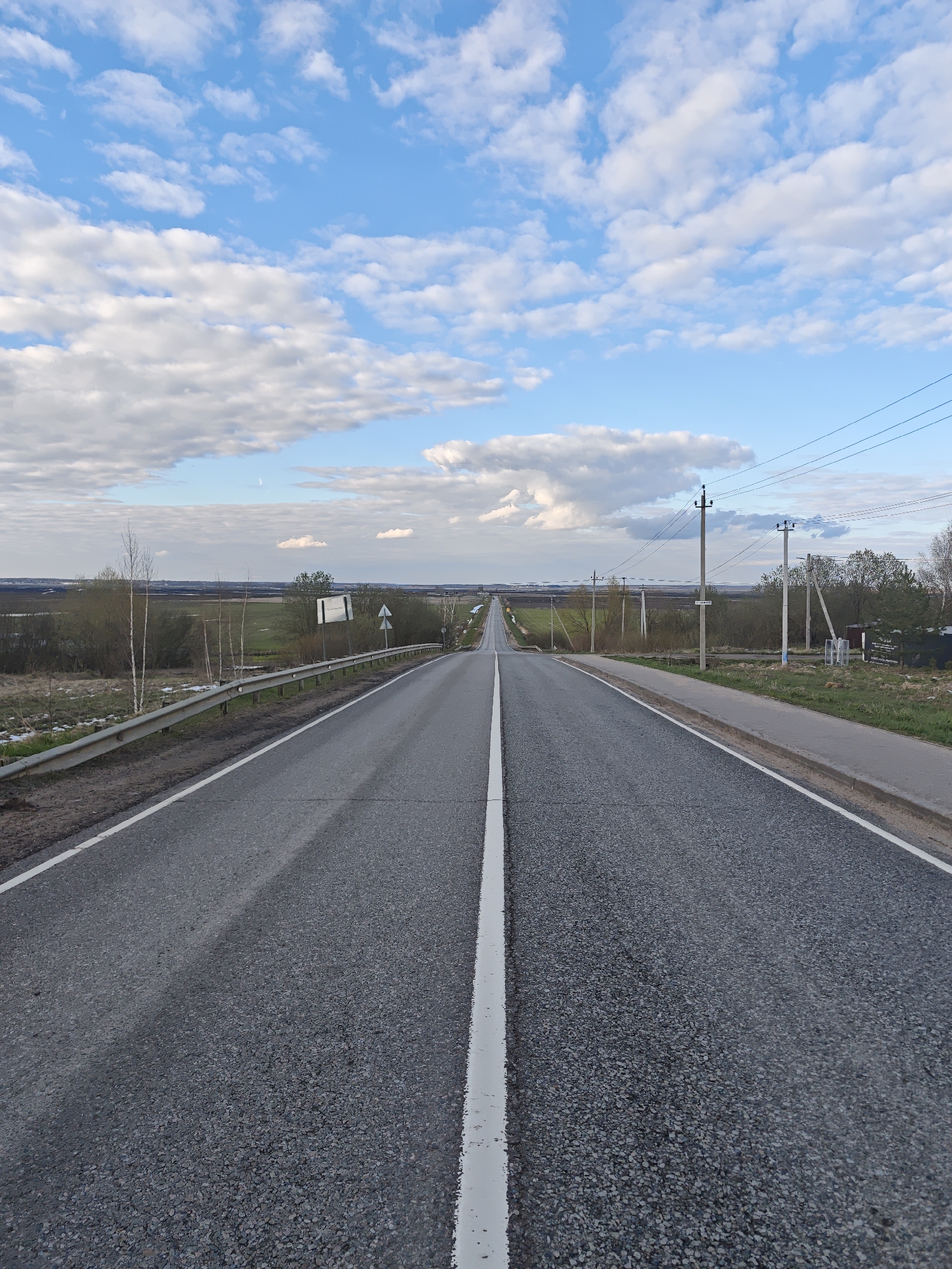 This image presents a wide view of a long, straight asphalt road extending into the distance under a vast sky, likely in Novosinkovo, Russia. The road, featuring a prominent white dividing line down its center and a solid white line along its right shoulder, appears to be a rural or intercity thoroughfare. The asphalt surface is dark grey with a visible gravel texture.
On the left side of the road, a metal guardrail runs parallel to the pavement. Beyond it, the landscape consists of green fields, some patches of lighter ground cover (possibly residual snow or dry grass), and several bare-branched trees, notably slender birches, and bushes. The terrain recedes into a flat, distant horizon.
To the right of the road, a narrow paved path or sidewalk runs alongside. Numerous utility poles with overhead power lines stretch into the horizon, mirroring the road's path. Further down, there are clusters of trees and a small, dark structure.
The sky is a bright blue, filled with scattered white, fluffy cumulus clouds, becoming denser towards the lower part of the sky. A faint crescent moon is discernible in the upper-left sky quadrant.
The time of day appears to be late afternoon or early evening, given the soft, diffused light and the visible moon. The weather is clear with moderate cloud cover. Several road signs are visible, including one on the left near the guardrail and others attached to utility poles on the right, but any text on them is illegible due to distance. The scene is devoid of people or explicit activity, conveying a sense of open space and tranquility.