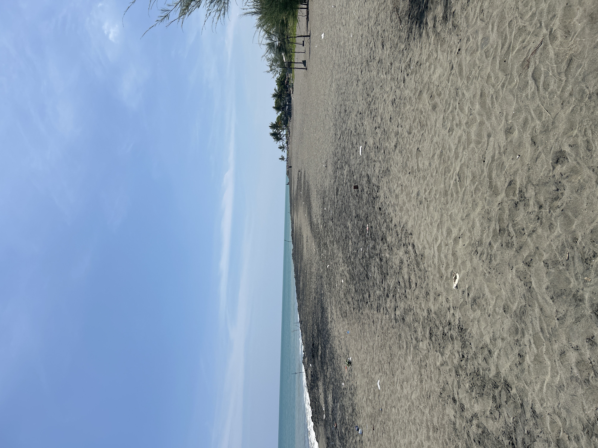 The image depicts a wide, sandy beach with dark gray sand, characteristic of the beaches along the coast of Banda Aceh, Indonesia. The ocean is visible on the left side, with gentle waves rolling onto the shore. The sky is clear and blue with scattered light clouds. The beach appears somewhat littered with small pieces of debris, suggesting it may be a less-maintained area or has recently experienced high tides. In the distance, a cluster of palm trees and some small structures are visible, hinting at a settlement or recreational area. A few people are sparsely scattered along the shoreline, and some rudimentary wooden structures, possibly makeshift shelters or seating, are present on the right side of the frame. The overall impression is of a natural, coastal environment that shows signs of human activity and presence, as well as some environmental impact from litter.
