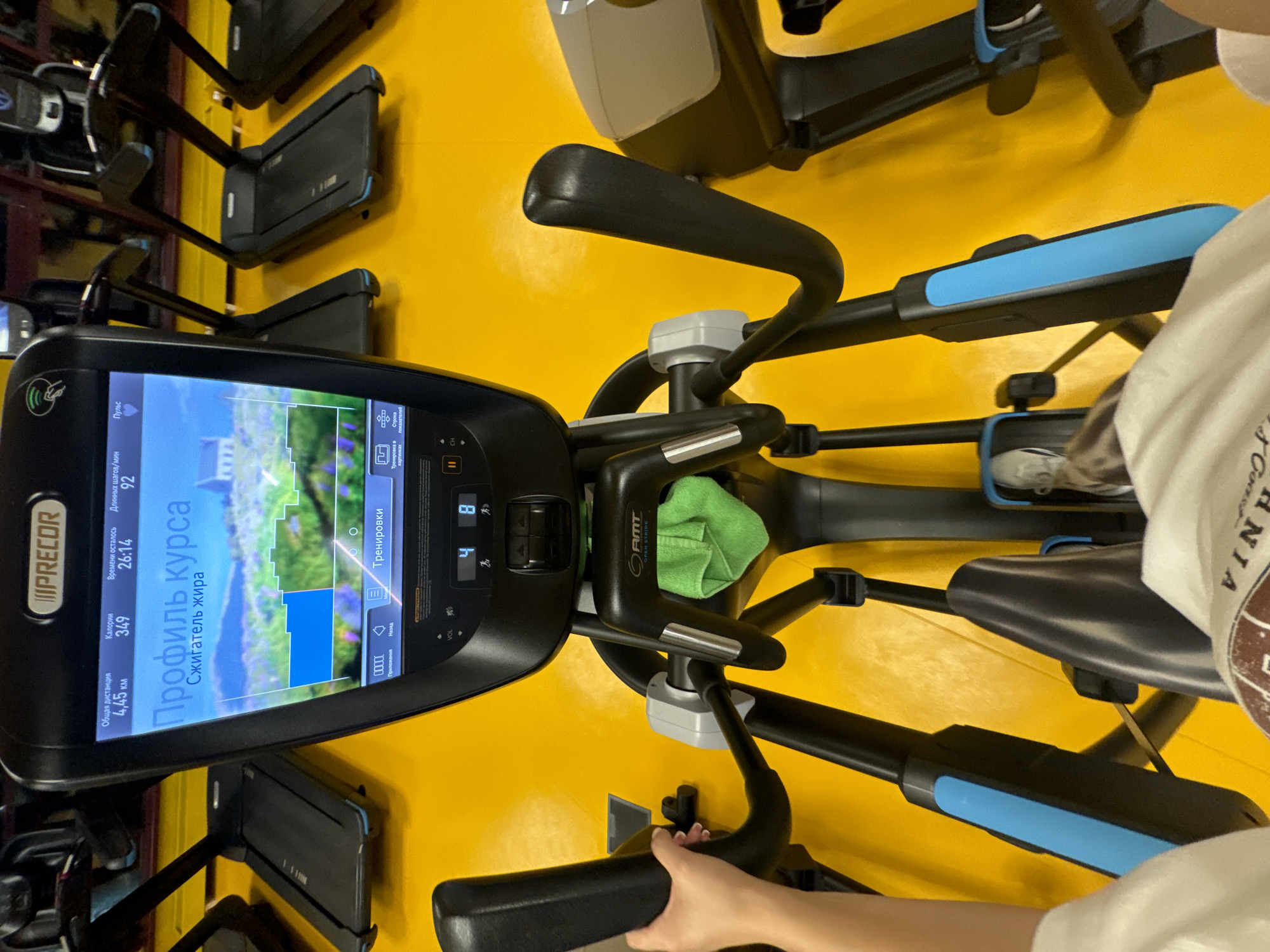 Here is a detailed description of the image:
**General Scene:** The image appears to be taken in a gym or fitness center in Vniissok, Russia, potentially indoors. The color scheme is predominantly yellow, with various pieces of gym equipment visible.
**People:** There are two individuals partially visible in the image. One person is on the right side of the frame, wearing a white shirt with some text visible on it. The other person is at the bottom of the frame. Their hands are holding onto the handles of an elliptical machine, suggesting they may be exercising.
**Subjects/Objects:**
* Elliptical machine: A large black elliptical machine dominates the central view of the image. It has a digital screen that shows data such as calories burned, remaining time, distance, pulse, and training profiles in Russian (e.g., "Профиль курса Сжигатель жира").
* Treadmills: In the background, there are black treadmills lined up against a wall.
* Towel: A green towel is draped over the center part of the machine.
* Other gym equipment: Other pieces of gym equipment are also visible.
**Actions/Interactions:** The person is seen holding the handles of the elliptical machine, indicating an action of using the exercise equipment.
**Environment:** The background has a solid yellow surface, which is likely the flooring of the gym. It appears to be a well-lit indoor environment.