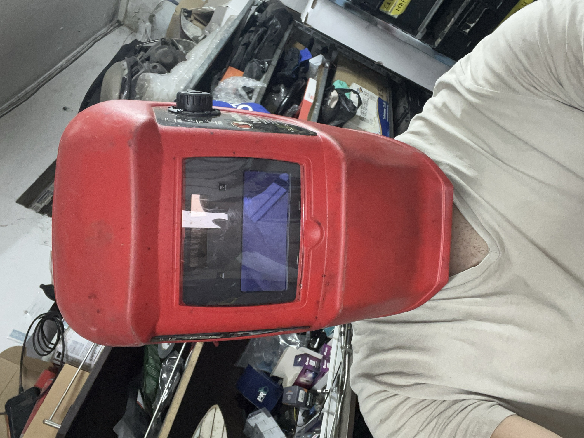 A first-person perspective selfie shows a person wearing a red welding mask and a light-colored t-shirt. The mask covers the entire face and extends over the top of the head. The t-shirt has a V-neck and visible chest hair. The background is cluttered with shelves filled with various items, including boxes, tools, and plastic packaging. The overall environment suggests a workshop or storage area, possibly in an industrial setting. The specific location within Burgas, Bulgaria, cannot be determined from the visual information provided.