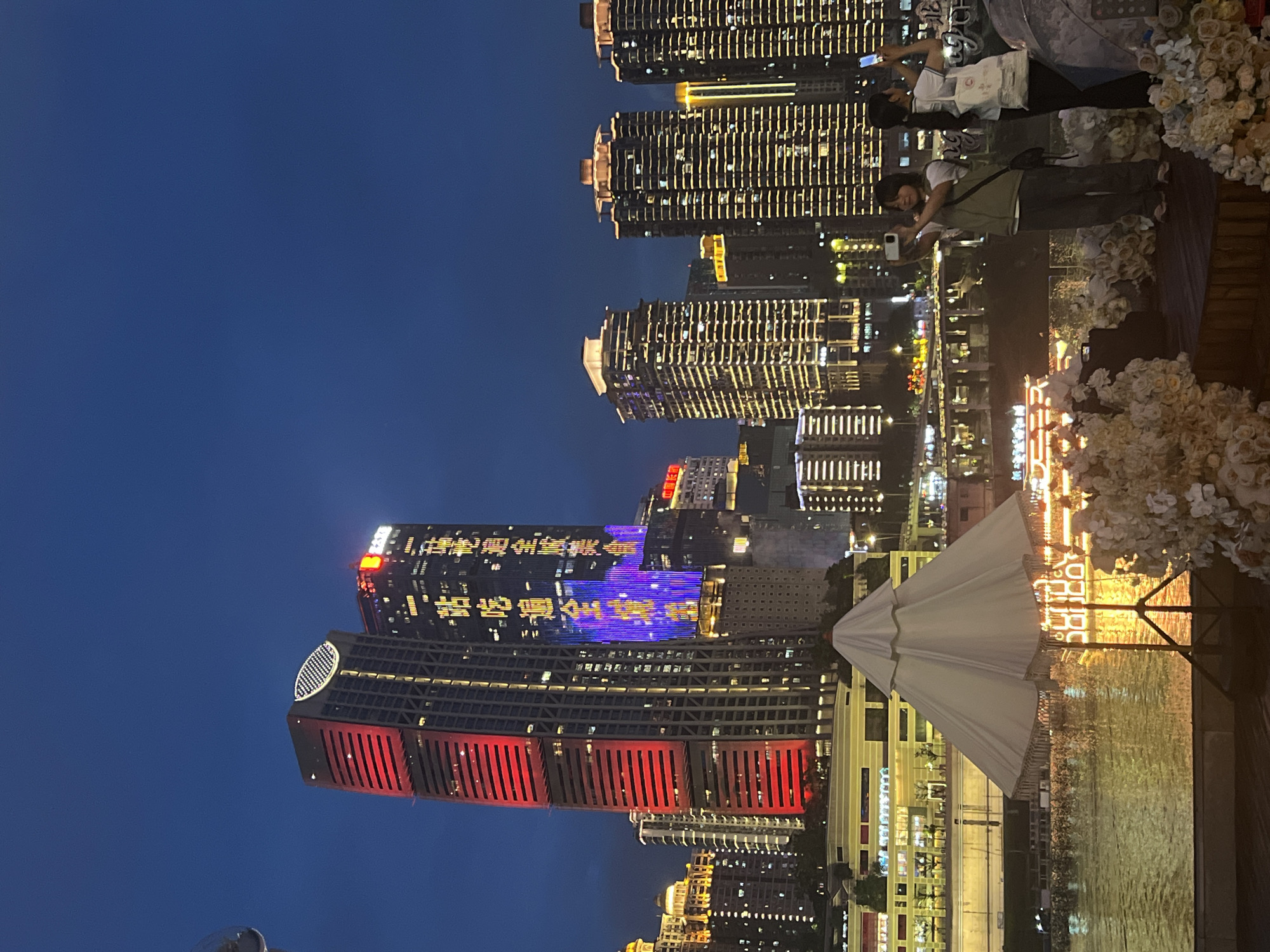 The image depicts a nighttime cityscape in Jiangbeicheng Subdistrict, China, dominated by illuminated skyscrapers and a waterfront area. Two young individuals are visible in the foreground, engaged in taking photos with their smartphones, suggesting they are tourists or enjoying a night out. They are positioned near a white umbrella and floral decorations, possibly part of a cafe, restaurant, or event space overlooking the river.
The buildings are a prominent feature, with many windows lit, creating a dazzling urban display. One skyscraper in the mid-ground displays a vibrant light show with what appears to be Chinese characters in yellow and blue, indicating a celebratory or promotional event. Another building in the foreground is characterized by a striking design featuring red vertical slats. The river or body of water in the foreground reflects the city lights, adding to the picturesque scene.
The time of day is clearly night, with a deep blue sky visible above the buildings. The weather appears to be clear, as the lights are sharp and reflected well on the water. The scene conveys a sense of lively urban energy and evening leisure. The visible text on the skyscraper appears to be Chinese characters, though the specific message is not fully decipherable from the image.