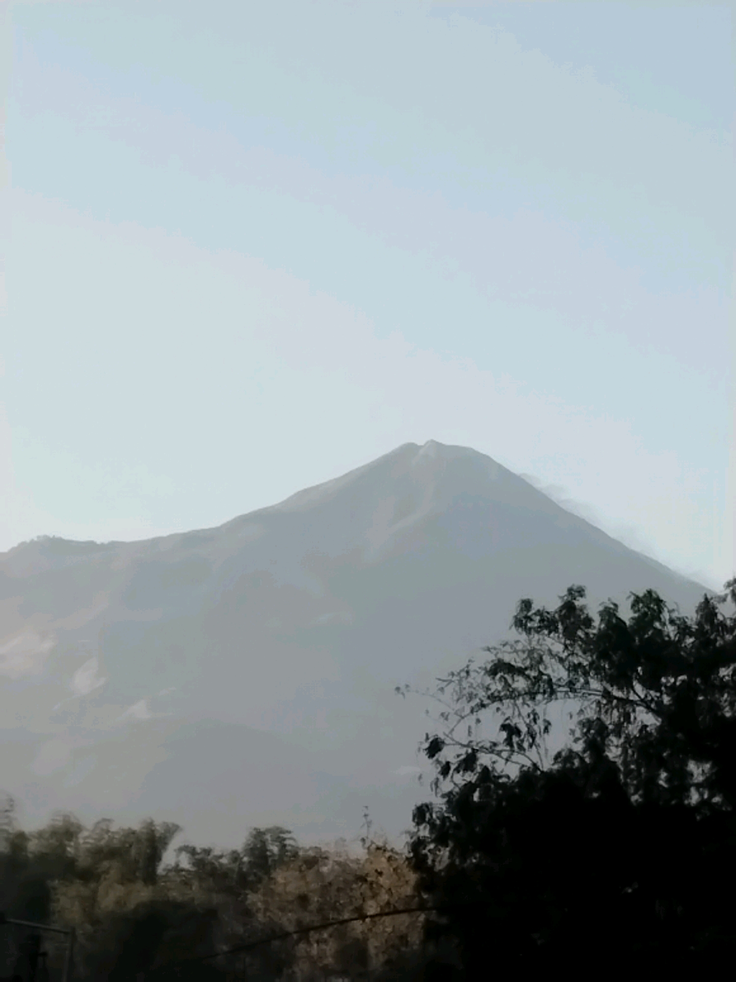 The image shows a hazy view of a mountain, likely Mount Merapi, in the distance. In the foreground, the silhouettes of trees are visible against a pale sky. The overall impression is one of a serene, possibly early morning or late afternoon scene, with the natural landscape dominating the frame. The location is identified as Kuripansari, Indonesia, a region known for its proximity to this active volcano.
