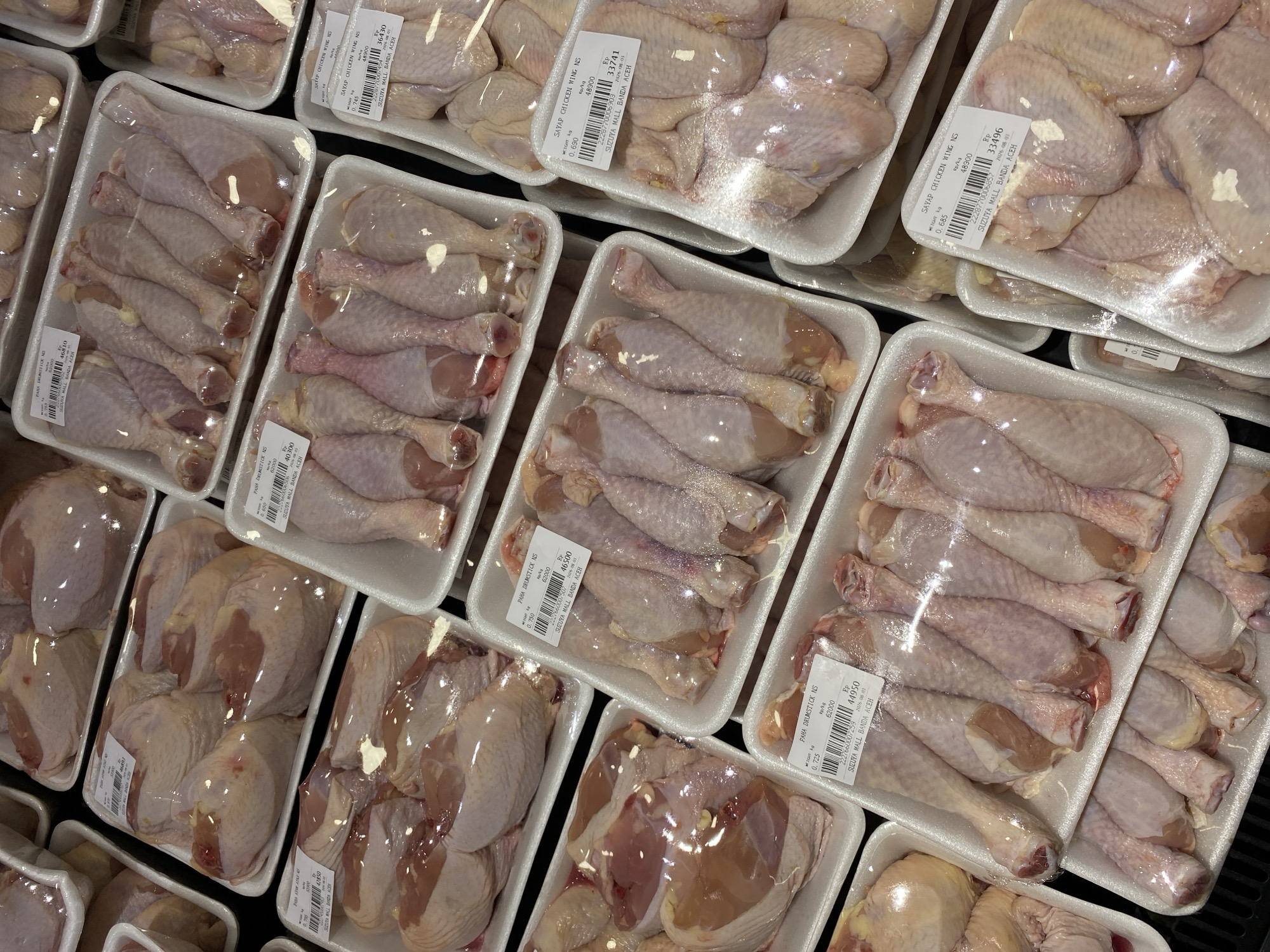 The image displays a close-up, overhead view of multiple white styrofoam trays filled with raw chicken parts, predominantly chicken drumsticks and wings, packaged in clear plastic. The trays are arranged in a grid-like pattern, suggesting they are likely on display in a market or grocery store. Some trays have visible price labels, with text that appears to be in Indonesian. One label explicitly mentions "SUZUYA MALL" and "BANDA ACEH", indicating the location as Suzuya Mall in Banda Aceh, Indonesia. The lighting appears to be artificial, common in indoor retail environments. There are no people visible in the image, only the products for sale. The chicken pieces are raw, with varying shades of pink and white meat, and some have visible skin. The overall scene is orderly and focused on the presentation of the food items.