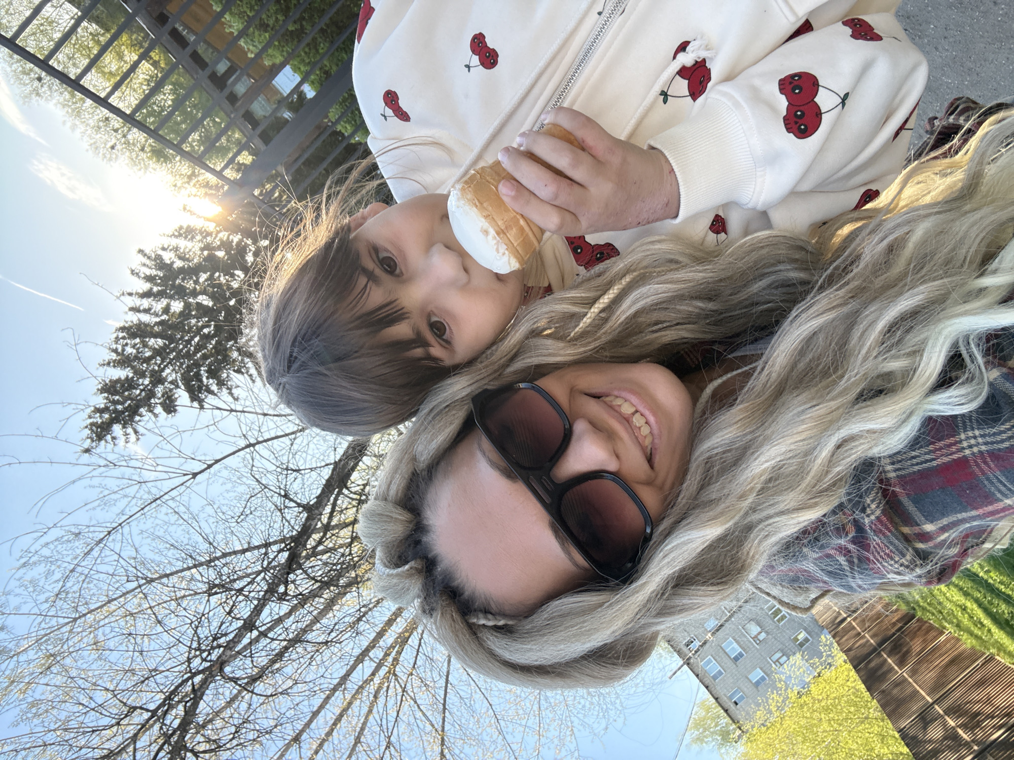 A selfie-style photograph, oriented sideways, shows a woman and a child outdoors in daylight. The woman is in the foreground, smiling, and wearing dark sunglasses and a plaid shirt. Her blond hair is styled in loose waves. Behind her and slightly above, a child holds an ice cream cone. The child wears a white jacket with red cherry patterns. The background includes bare trees, a blue sky, a building with windows, and what appears to be a wooden fence. The presence of trees and buildings suggests an urban park setting in Rusanovo, Russia.