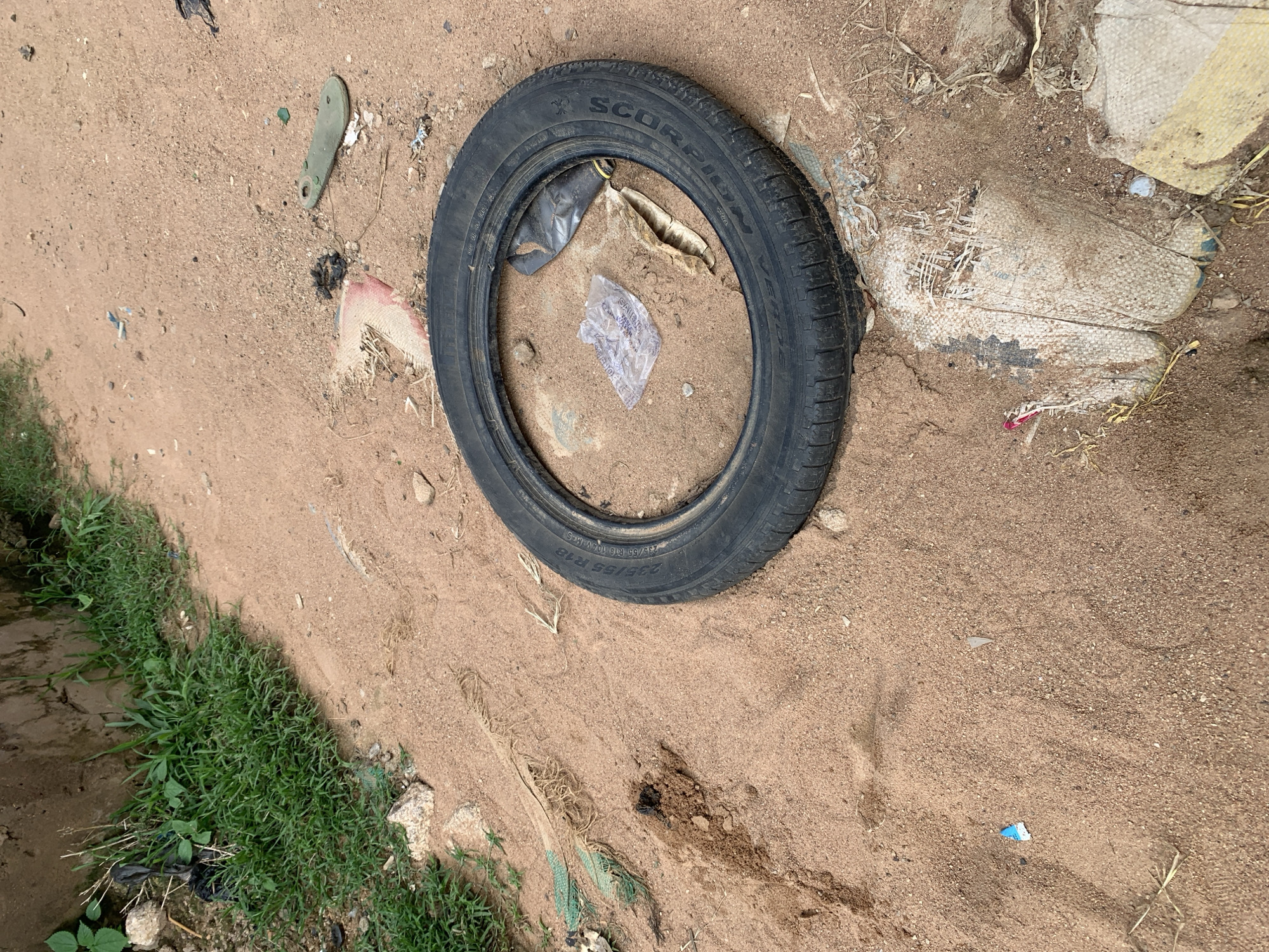 An overhead, eye-level shot shows a used car tire lying flat on the ground in a patch of dry, sandy soil. The tire is black and has "SCORPION VERDE" and "235/55 R18 104V M+S" imprinted on its sidewall. Inside the tire, there is a crumpled piece of clear plastic packaging and a dried leaf. To the left of the tire, a small pile of dark earth is visible, with some green vegetation growing from it. Further left, the edge of a rough, gray concrete wall is present, with more green grass growing along its base. To the right of the tire, pieces of torn, beige woven fabric are partially buried in the sand. A light green, oval-shaped object with a hole in the middle lies in the sand to the upper right of the tire. A few small pieces of blue and white plastic are scattered in the sand to the lower left. The overall scene suggests a neglected outdoor environment with scattered debris.