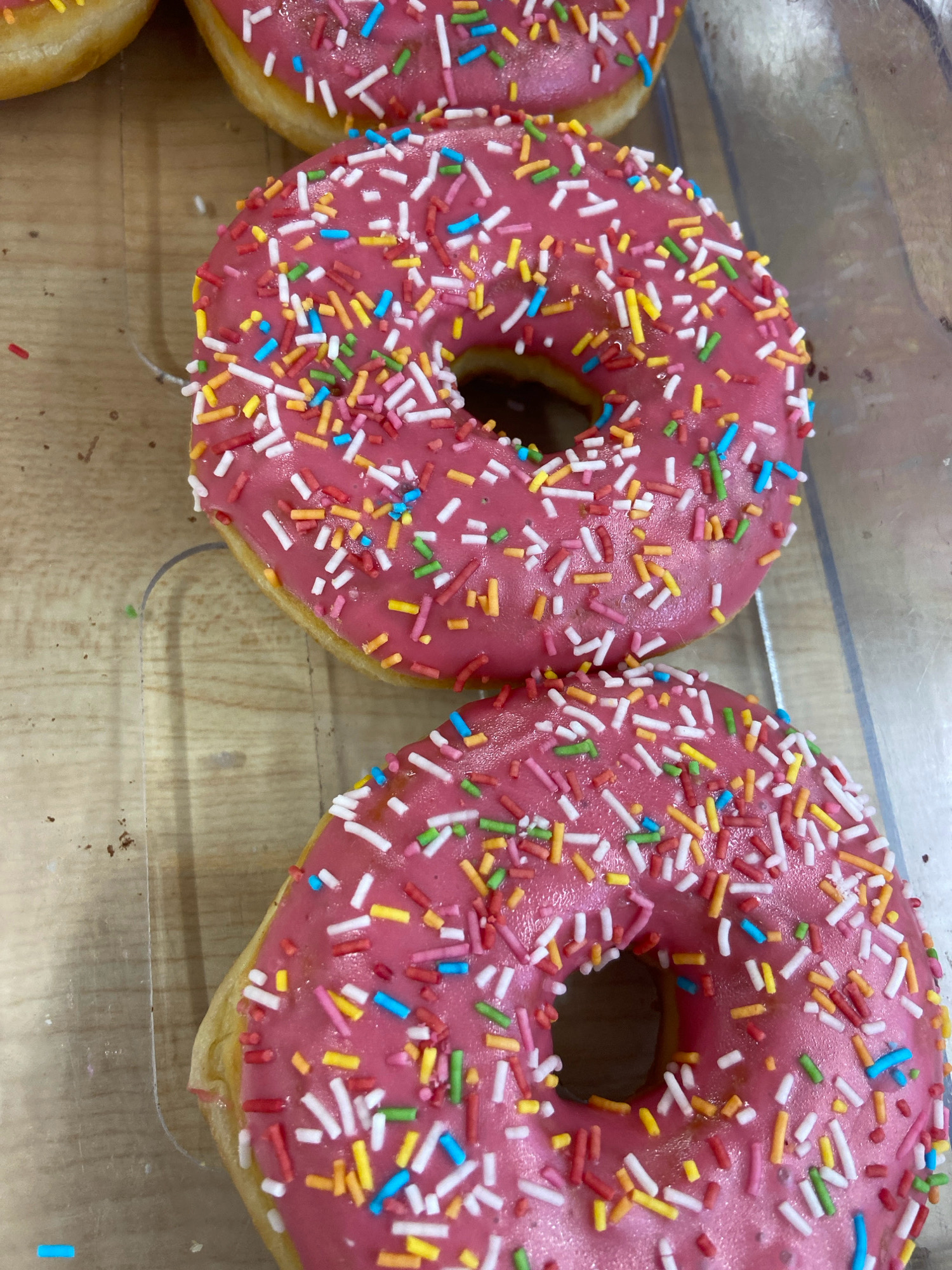 A close-up, overhead shot displays two pink frosted donuts with colorful sprinkles arranged diagonally on a light brown, textured surface, possibly a wooden counter or tray. The donuts are positioned one above the other, slightly offset, creating a sense of depth. The top donut is partially obscured by the bottom one, and a small portion of another donut with yellow frosting is visible at the top left corner. The donuts are placed within a clear plastic container, suggested by the visible rim and a slight glare on the right side. The overall lighting is bright, highlighting the textures and vibrant colors of the sprinkles and frosting. This scene is consistent with a bakery or confectionery display in Balashikha, Russia, where such pastries are commonly sold.