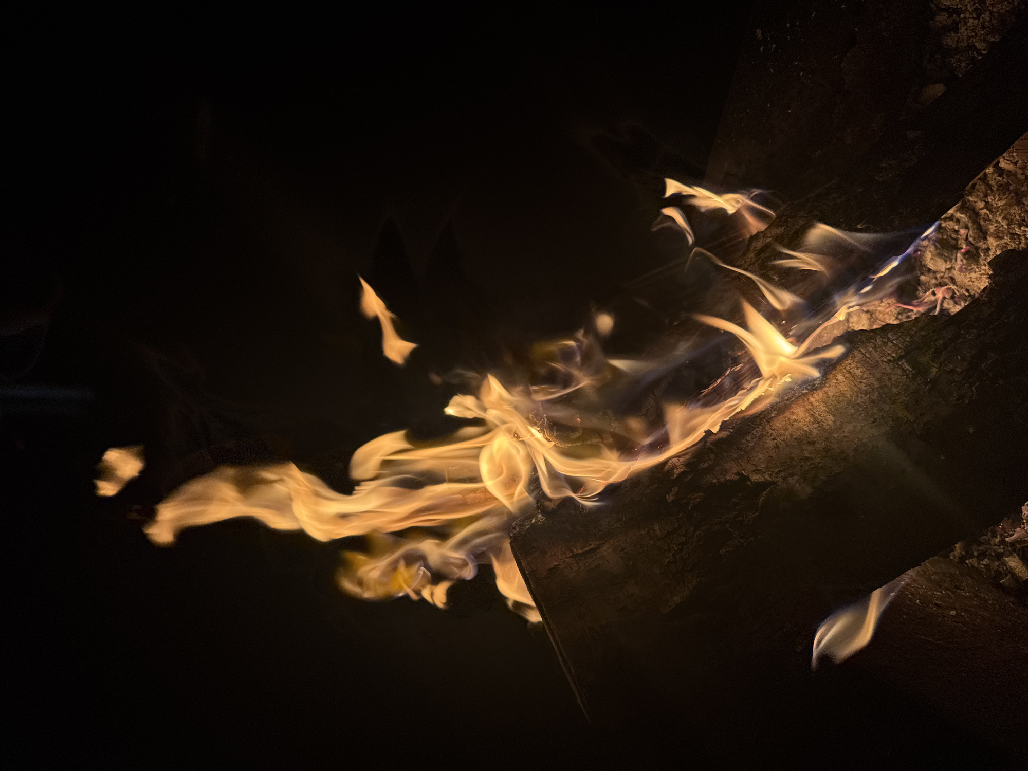 The image depicts a close-up view of flames engulfing a piece of wood. The fire is the primary focus, with orange and yellow hues contrasting against the dark background. The wood appears to be burning intensely, with details of its texture and charred surface visible. The black background dominates the scene, creating a high contrast that accentuates the flames. There are no people or other objects present. The absence of contextual elements suggests that this is an isolated shot of a fire. The location in the city of Mayak, Russia is not evident from the image.