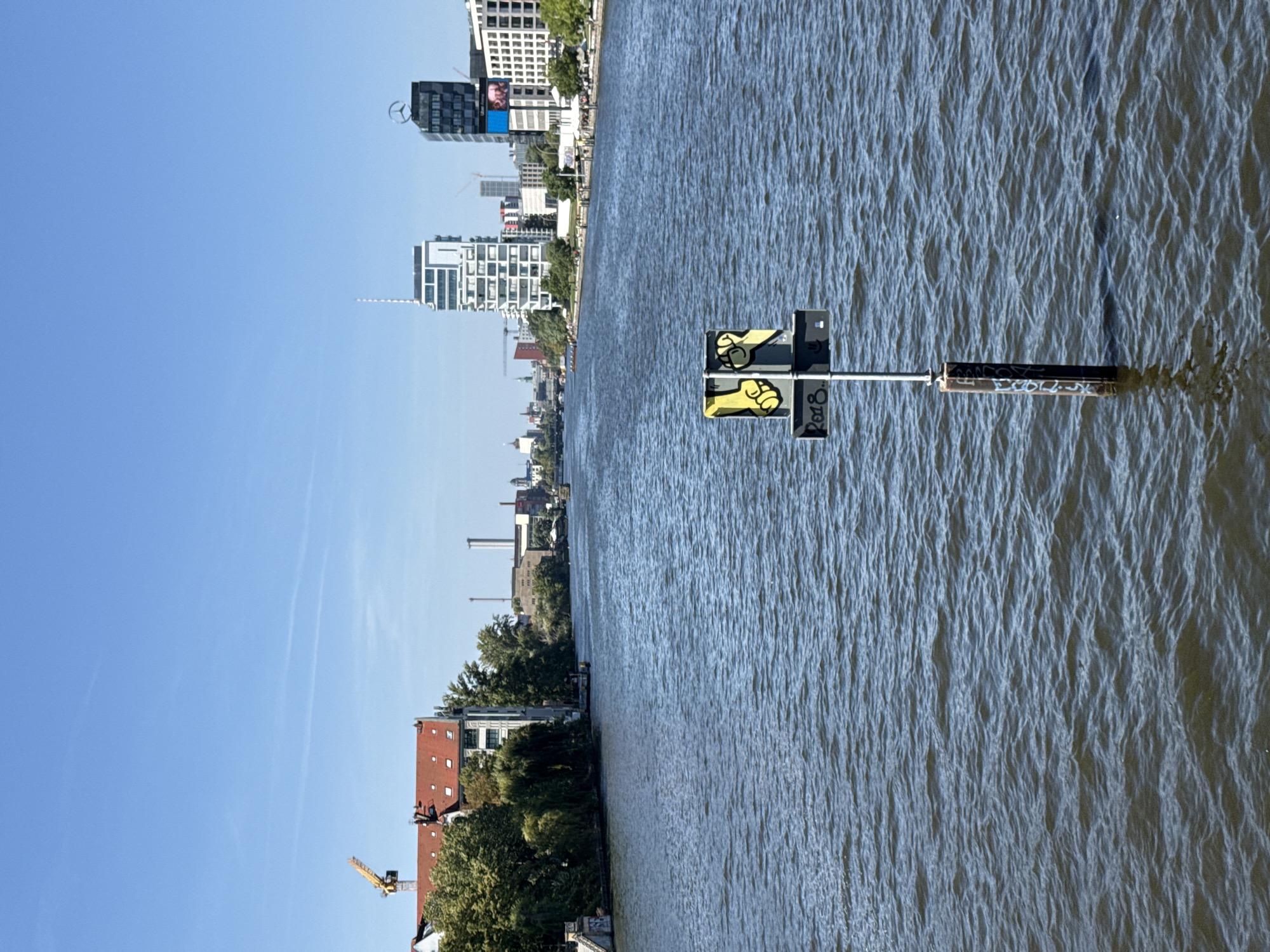 The image depicts a wide, calm river flowing through an urban environment under a clear blue sky. In the foreground, a metal post rises from the water, supporting a navigational sign with a graphic of two raised yellow fists and the text "2018" below. The riverbanks are lined with buildings, trees, and infrastructure.
On the left bank, a red-roofed building with multiple windows is visible, partially obscured by lush green trees. A yellow construction crane stands behind this building. Further along the river, industrial-looking structures with tall smokestacks are present.
On the right bank, a modern, white, multi-story building with a distinctive geometric facade dominates the skyline. Next to it stands a dark building topped with a Mercedes-Benz logo. Other buildings of various architectural styles, including a white building with many windows, are scattered along this bank. People can be seen on the right bank, some near tables and umbrellas, suggesting an outdoor seating area. Boats are also visible on the water in the distance. The overall impression is that of a busy, contemporary cityscape integrated with a natural waterway, likely Berlin, Germany, given the architectural styles and the presence of the Mercedes-Benz logo on a building.