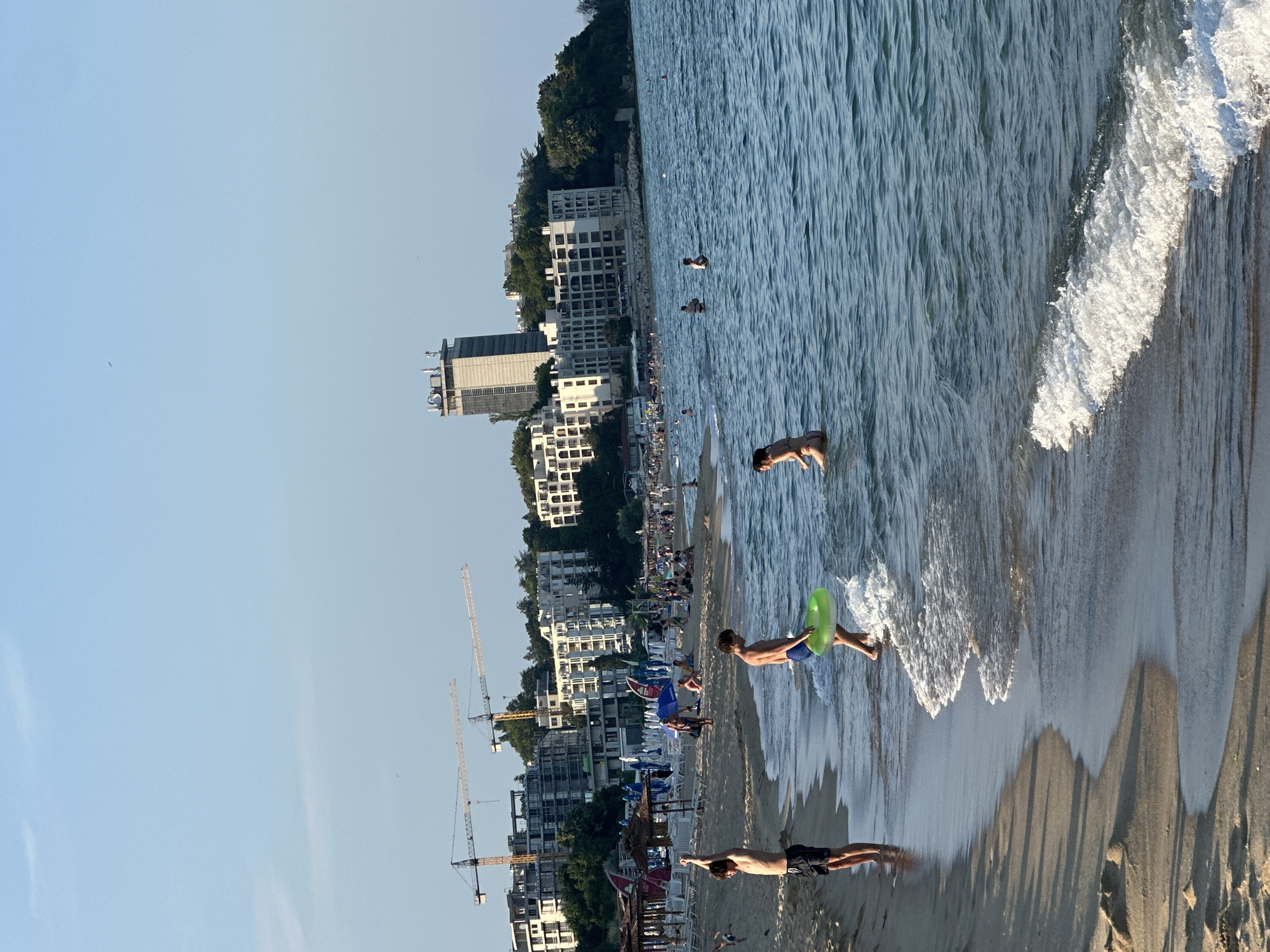 The image depicts a crowded beach scene with people enjoying the water and the sand. In the foreground, two young boys are in the shallow water, one with a green inflatable ring. A third boy stands on the wet sand, stretching his arms. Behind them, a busy beach is lined with umbrellas and lounge chairs, with many people scattered across the sand.
In the background, a coastline is visible with several large buildings, including a tall, modern skyscraper. Construction cranes are also prominent, suggesting ongoing development in the area. The sea is a calm blue, with small waves breaking on the shore. The sky is clear and blue. The overall impression is of a vibrant, sunny day at a popular seaside resort.