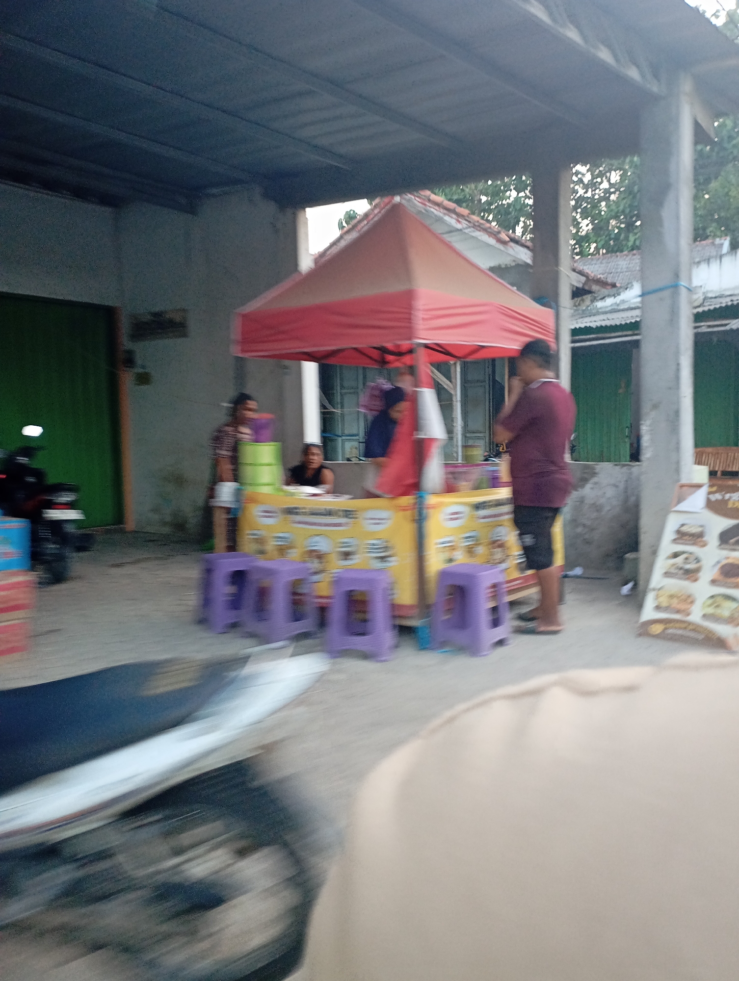In the city of Kecamatan Haurgeulis, Indonesia, a food stall operates under a red and orange awning. The stall displays a yellow banner with food images and text. Several purple plastic stools are arranged in front of the stall. A man in a maroon shirt and black shorts stands to the right of the stall, holding his phone to his ear. To the left, a woman in a striped shirt stands behind the stall, and another person is partially visible. A motorcycle is parked to the far left, and boxes are stacked nearby. In the background, buildings with green doors and tiled roofs are visible. The scene appears to be outdoors, with trees and foliage in the distance.