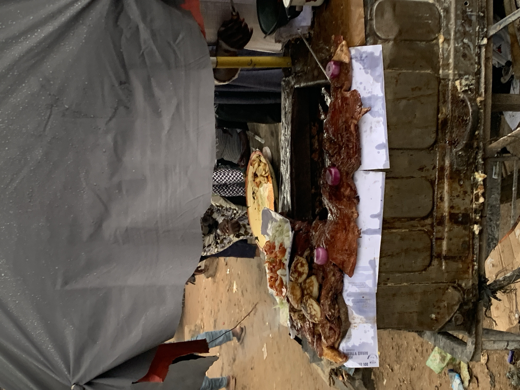 The image depicts an outdoor food stall in Rafin Par, Nigeria. A variety of grilled meats, including what appears to be steak and possibly chicken, are prominently displayed on a makeshift counter. These meats are seasoned and some are garnished with slices of red onion. Alongside the meat, a plate of chopped tomatoes and shredded lettuce is visible, suggesting a salad component. A large, slightly weathered umbrella or tarp provides shade over the stall. In the background, people can be seen milling about in a dusty, unpaved area, characteristic of a marketplace or street scene. The stall itself appears to be constructed from metal, with a grilling surface visible beneath the food. The overall impression is of a bustling street food vendor in a Nigerian town.
