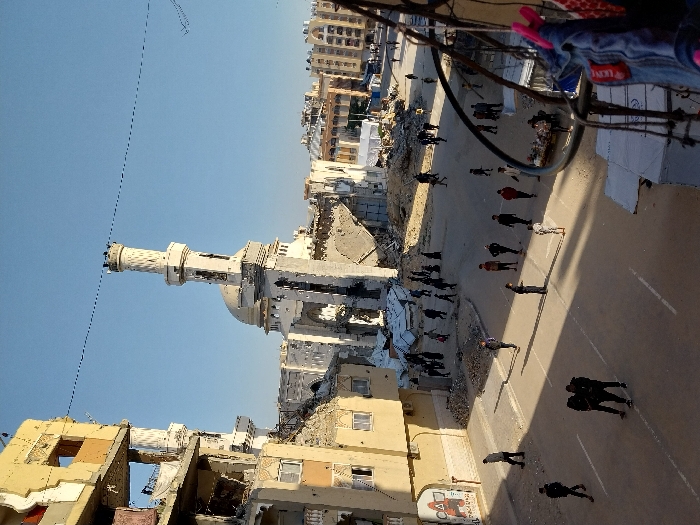This image depicts a street scene in Khan Yunis, Palestinian Territory, characterized by extensive structural damage under a clear blue sky.
In the mid-ground, a large mosque stands partially destroyed, its dome visible, and one minaret largely intact but leaning slightly. Surrounding sections of the mosque are collapsed, exposing rubble and skeletal structural elements. To the left, multi-story residential or commercial buildings are severely damaged, with entire sections of walls missing, revealing interiors, exposed rebar, and debris. One such building on the left features graffiti on its lower wall, including a red triangular symbol.
The paved street in the foreground and mid-ground shows white dashed road markings. Numerous individuals, estimated to be between 30 and 40, are distributed across the street and areas covered in rubble. They are observed walking, standing, and navigating the damaged environment. Attire varies, with some individuals in darker clothing and some women wearing head coverings. In the mid-ground right, one person appears to be operating a small street stall or cart.
Piles of concrete, twisted metal, and other debris are scattered around the damaged structures and on parts of the ground. In the far background, additional buildings, some appearing less damaged, are visible. Overhead, thin black wires traverse the upper portion of the frame. The photograph appears to be taken from an elevated vantage point, with a dark, curved railing and what looks like a denim garment hanging on it visible in the extreme foreground on the right. Strong sunlight casts distinct shadows across the street and buildings, indicating a clear day.