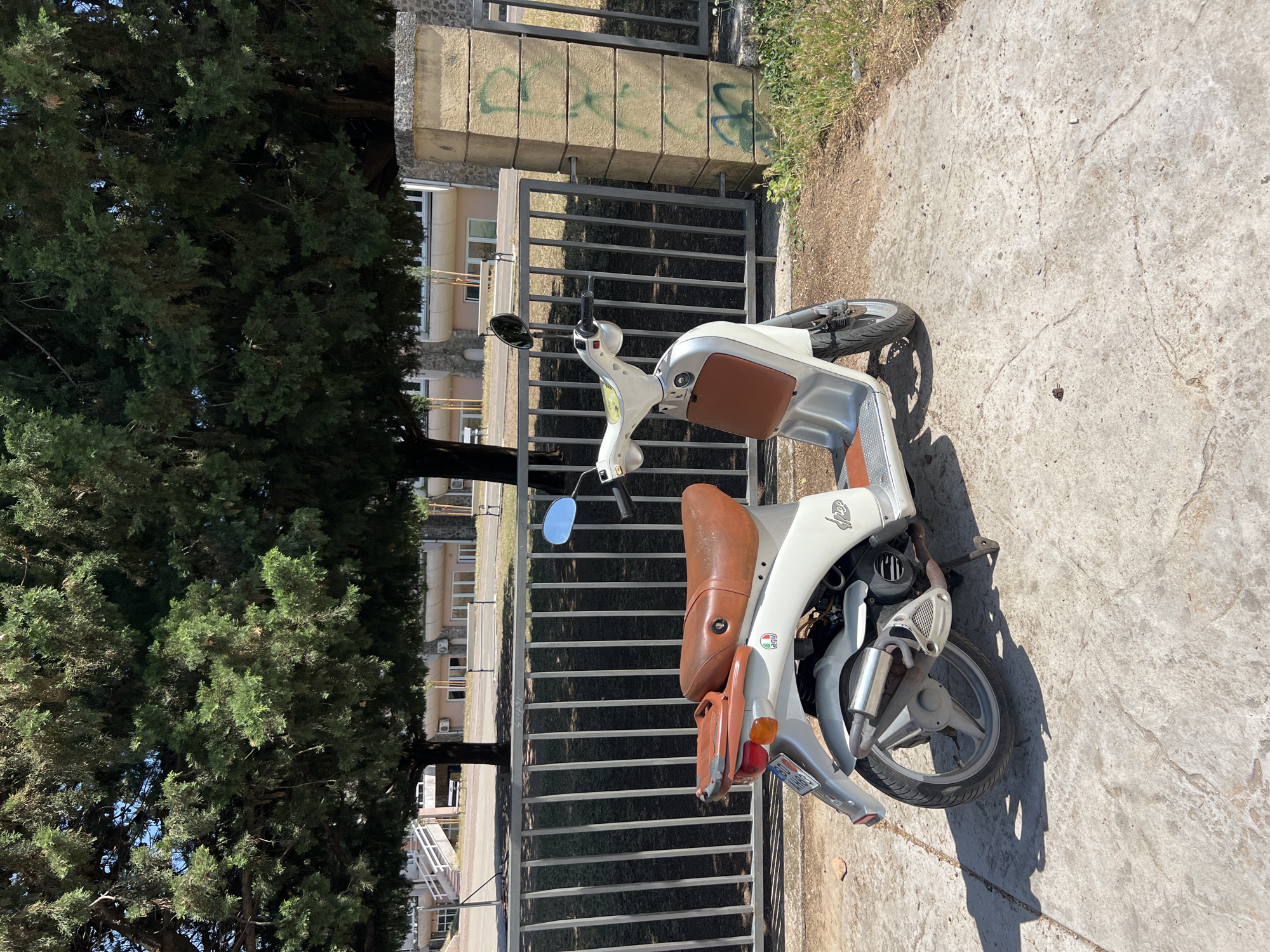 A white and brown scooter is parked on a concrete walkway in front of a metal fence. Behind the fence is a paved area with some buildings and trees. The scooter is a Piaggio Free, with a brown seat and floorboard accents. The scooter has a license plate that reads "BD 080 AE". The scene is outdoors in what appears to be a public space, possibly a park or school grounds. The scooter is stationary and there are no people in the image. The lighting suggests it is daytime.
