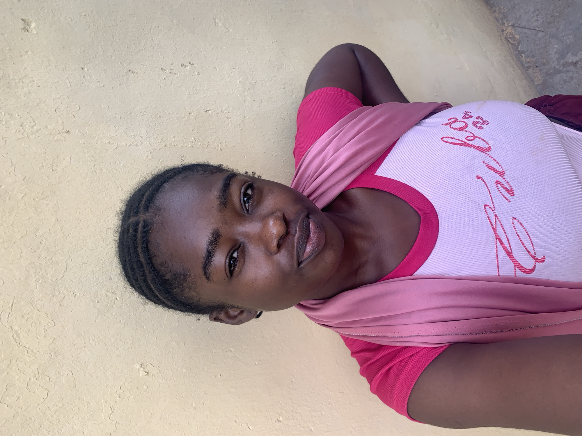 A medium shot presents a Black woman leaning against a light-yellow wall. Her hair is styled in tight braids. She is wearing a white, ribbed top with a pink collar and pink writing across the front, overlaid by a pink shawl or scarf. The location is Rafin Par, Nigeria. The immediate environment is a simple wall with a neutral coloration. The image provides minimal context beyond the subject and the wall.