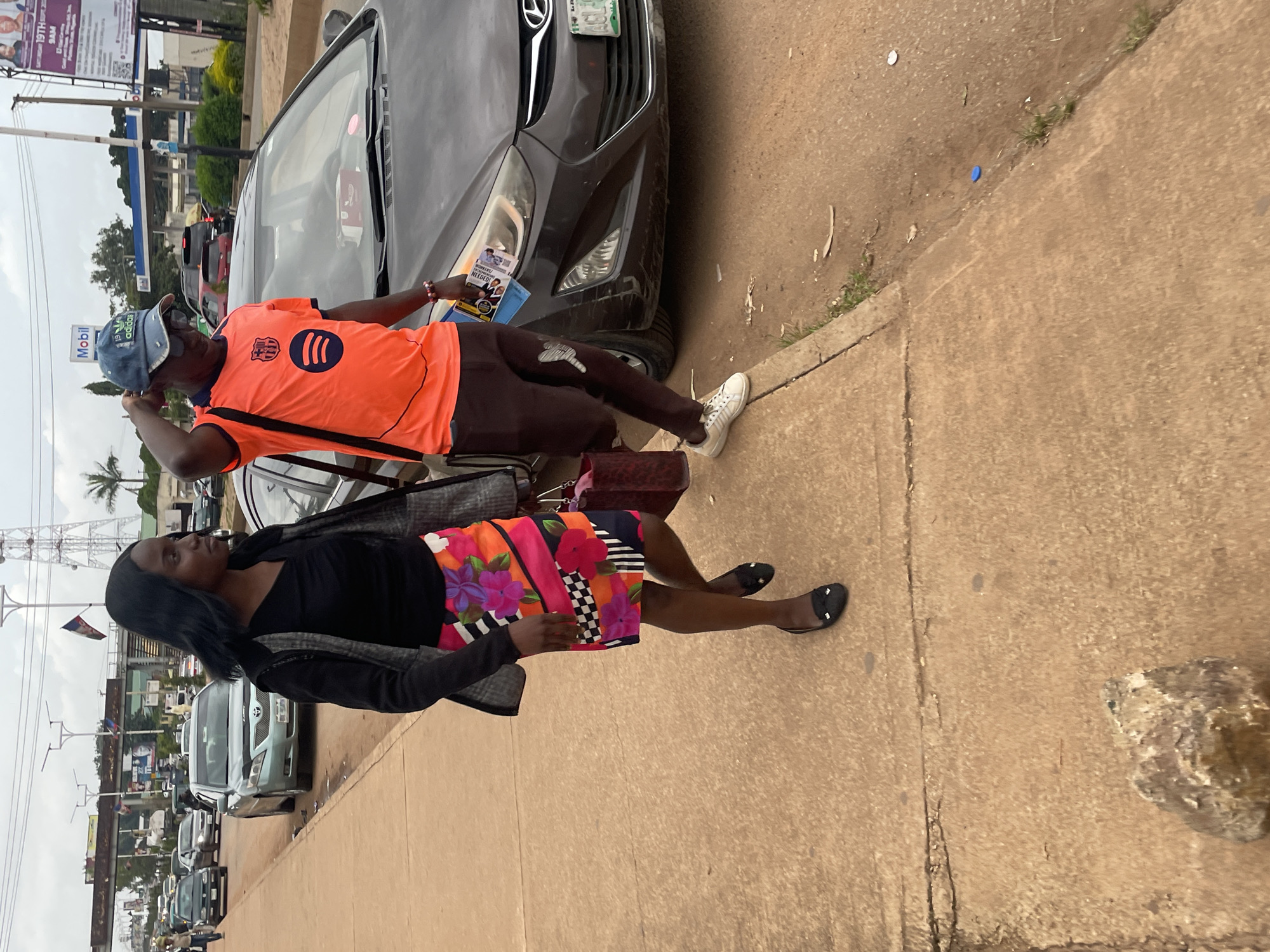 Two individuals, a woman and a man, walk along a paved sidewalk next to a street with several cars. The woman, on the left, wears a black top, a vibrant floral print skirt, and black flats. She carries a red patterned bag. The man, on the right, is dressed in an orange t-shirt with a logo, dark pants with a white design, a blue baseball cap, and white sneakers. He holds a stack of papers. A dark-colored car is parked on the street to the right of the man. In the background, various vehicles are visible on the road, and a tall communication tower stands against an overcast sky. The scene appears to be in Jos, Nigeria, suggested by the "Mobil" gas station sign.