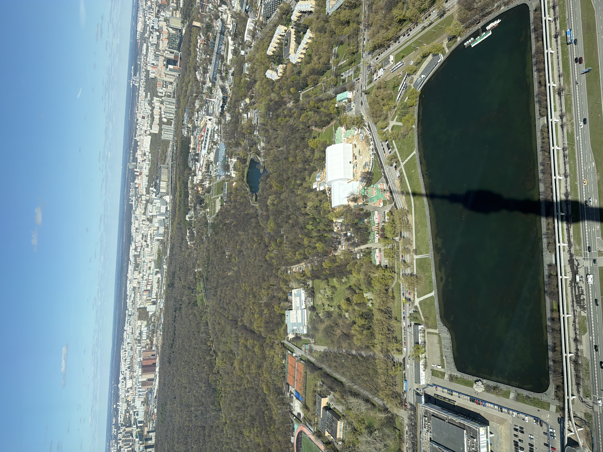An aerial view showcases a portion of Khimki, Russia, with a prominent body of water resembling a rectangular lake situated at the center-right. The lake is adjacent to a multilane highway, divided by a median and bordered by mature trees. A dense forest and green space cover a substantial area to the left of the lake, and within this green space, several buildings, including a large white structure and a smaller pond, are visible.
Beyond the green space, a cityscape extends into the distance. The city comprises numerous buildings of varying heights and architectural styles. The overall scene is captured under a blue sky, with light clouds scattered across the top-left. A shadow, likely from the observation tower, is cast over the lake.