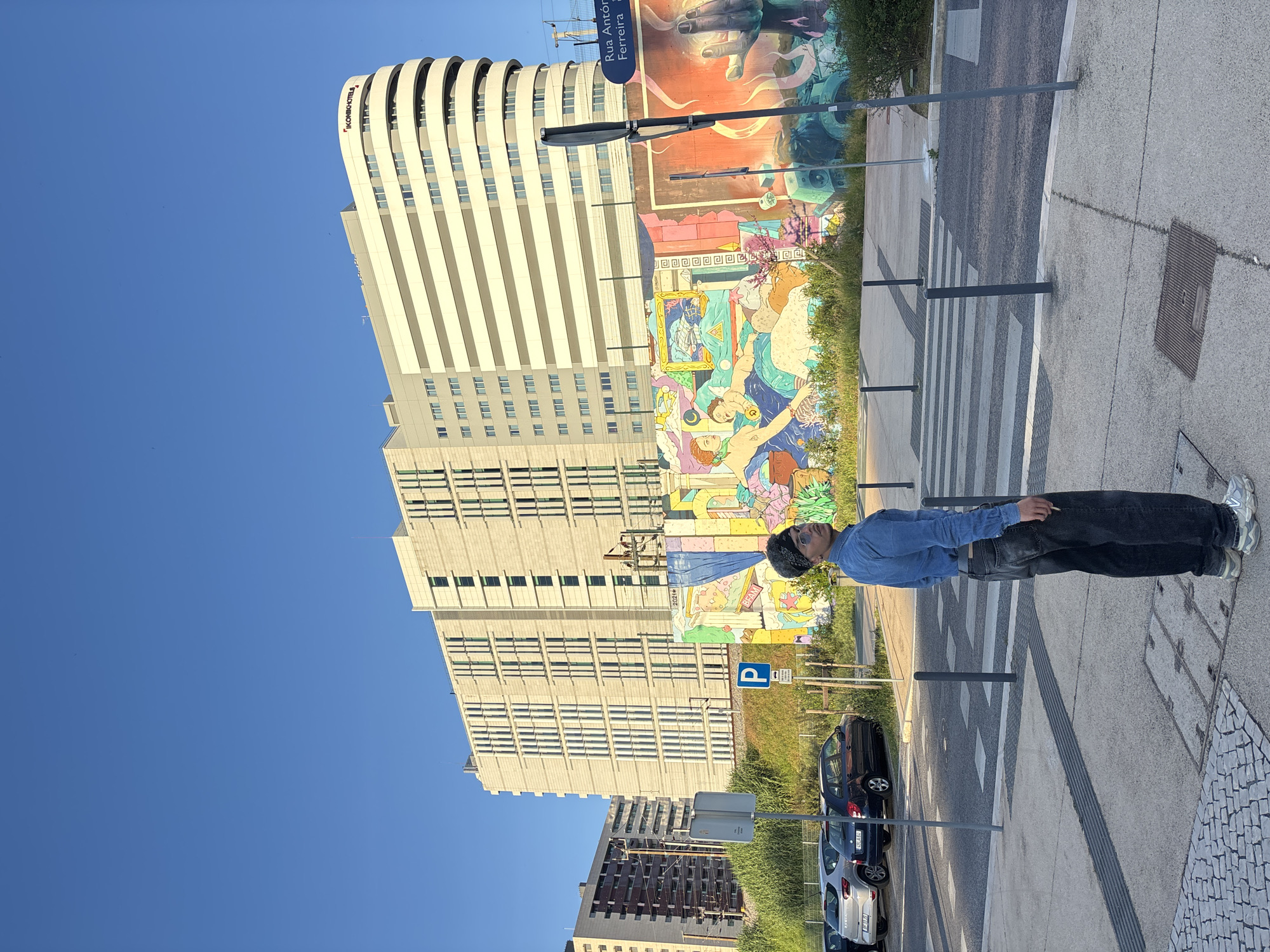 The image shows a man standing on a crosswalk in Lisbon, Portugal. He is wearing a blue denim shirt, black pants, and sneakers. He is also wearing a black beanie and sunglasses. He has dark hair. He is standing in front of a large mural on a building. In the background are other buildings, including the Ikonik Hotel Lisboa. Cars are parked along the street. The sky is blue. A sign reads "Rua António Ferreira". A parking sign with a blue "P" is visible.