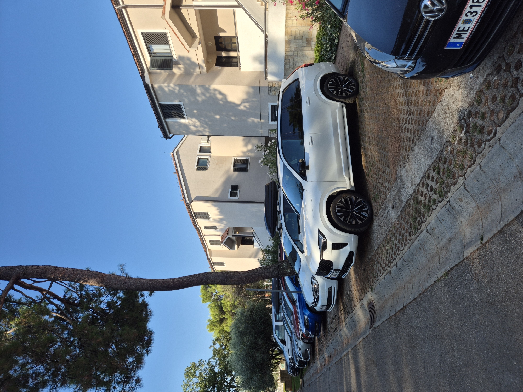 The image shows a row of parked cars in a residential area. The cars are parked on a paved area next to a road. There are several buildings in the background, which appear to be apartments or townhouses. The sky is clear and blue, and the sun is shining. The scene appears to be in Umag, Croatia, given the presence of a German license plate on one of the cars. The cars are as follows, from foreground to background: a black Volkswagen Golf, a white Volvo XC90, a white Volkswagen Passat, a blue Skoda Octavia, a silver Volkswagen Passat, and a white Volkswagen van. The white Volvo XC90 has a black roof box. There is a large tree in the foreground, and some other trees and vegetation in the background. The architecture of the buildings is typical of Mediterranean style, with tiled roofs and cream-colored walls. The cars are all facing in the same direction, suggesting they are parked in a designated parking area. The presence of the German license plate could indicate that the cars are owned by tourists or residents who are from Germany. The overall impression is one of a quiet, residential street on a sunny day.
