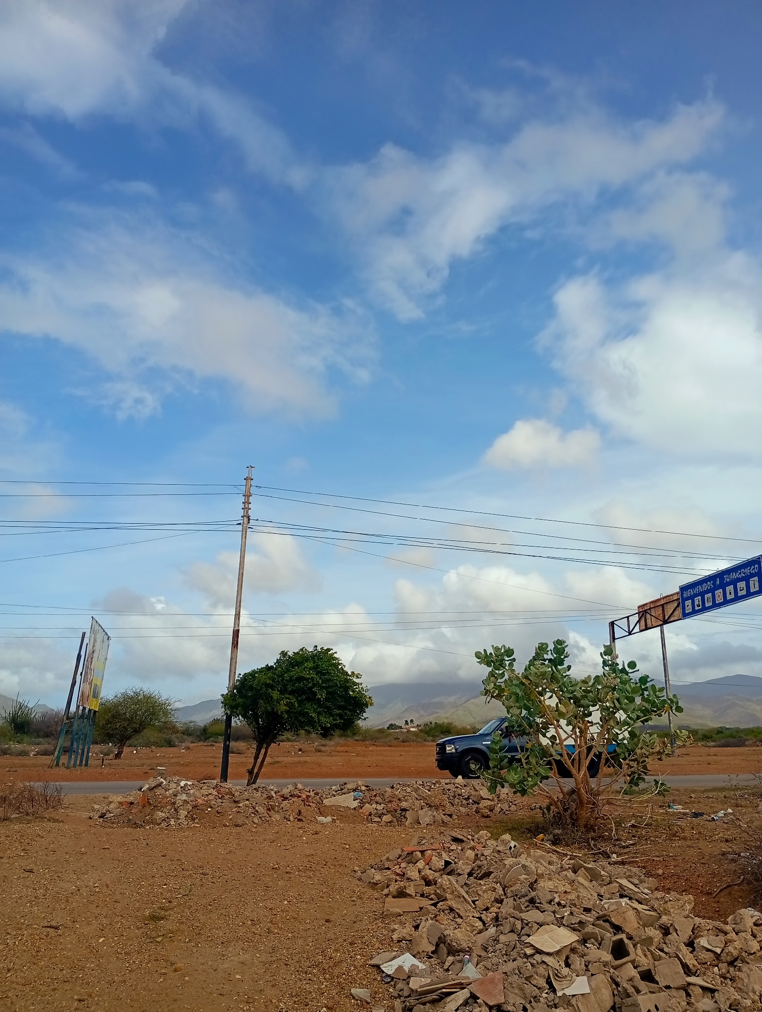 The image depicts a roadside scene in Altagracia, Venezuela. In the foreground, piles of rubble and debris are scattered across dry, reddish-brown soil. A sparse, thorny bush is visible on the right. Beyond the rubble, a paved road runs horizontally across the frame. A dark blue pickup truck is parked on the side of the road, partially obscured by a leafy green tree. Further back, a solitary utility pole with multiple wires stands tall against a bright blue sky dotted with white clouds. Two trees with green foliage are situated in the middle ground, one behind the utility pole and another to the left. A large, weathered billboard is leaning precariously on the left side of the image. On the right, a blue sign with white text and icons is suspended from a metal structure above the road, indicating "BIENVENIDOS A JUANGRIEGO" (Welcome to Juangriego). In the background, distant mountains are visible under a cloudy sky. The overall environment appears arid and rural. There are no people visible in the image.