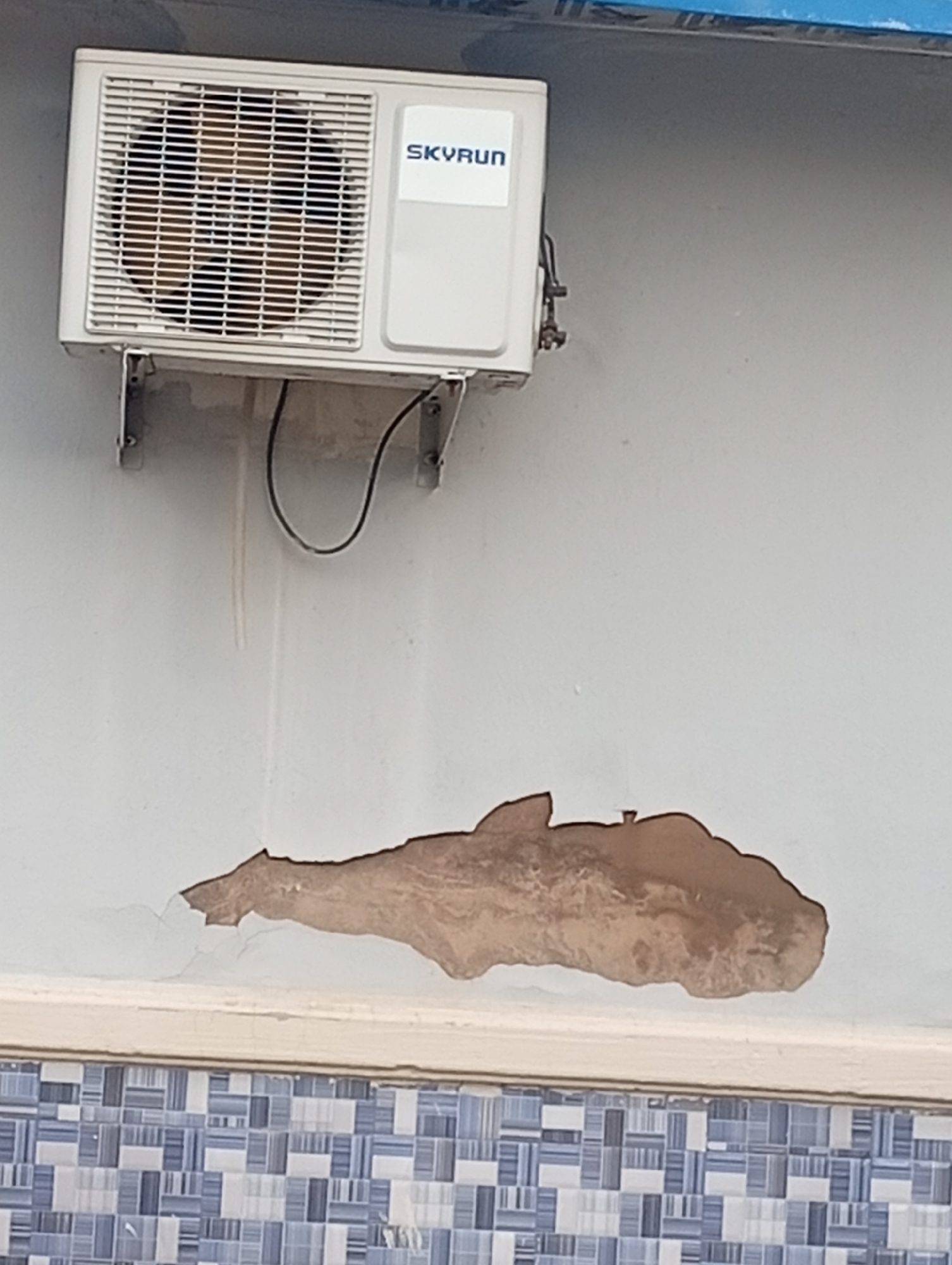 The image shows a close-up, eye-level shot of an outdoor wall with an air conditioning unit mounted on it. The wall is painted a light, muted color, and there is a noticeable patch of peeling paint, revealing a brown surface underneath. Below the peeling paint, there's a decorative tile border featuring a pattern of small, blue and white squares.
The air conditioning unit is white and mounted on metal brackets. It has a prominent grille on the front, and a logo that appears to say "SKYRUN" in blue lettering is visible on the right side of the unit. A black cable runs from the unit down the wall. The overall impression is of a somewhat weathered exterior surface.
There are no people visible in the image. The scene appears to be a section of a building's exterior. The lighting suggests it might be daytime, but there are no strong shadows or clear indicators of the time of day or weather conditions. The location context of Jalingo, Nigeria, implies a potentially warm climate. The peeling paint and visible wear suggest this is an outdoor environment that has been exposed to the elements. No specific activity or event is taking place; it is a static scene focused on the wall and the air conditioning unit.