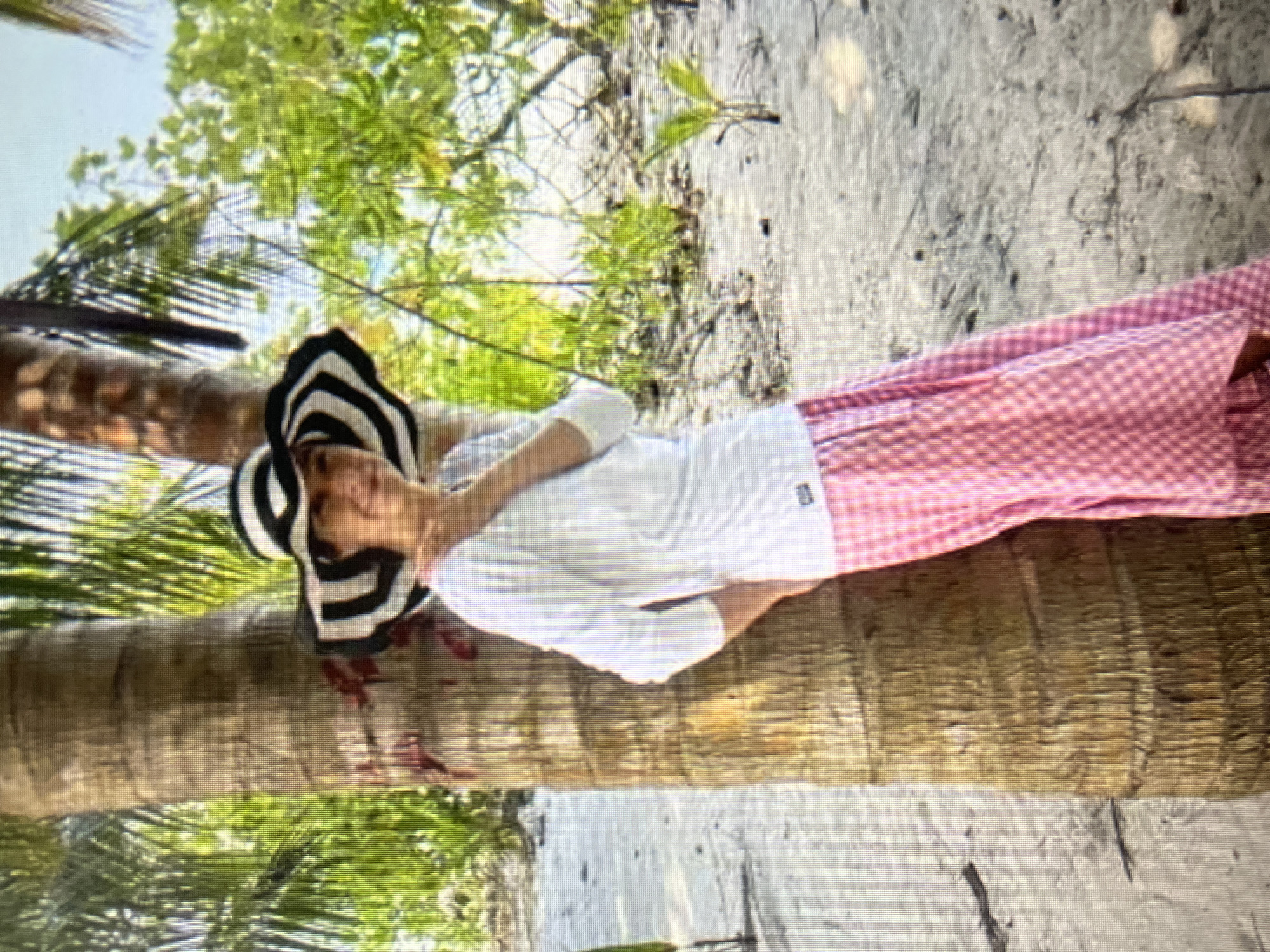 The image shows a woman wearing a wide-brimmed black and white striped hat, a white long-sleeved shirt, and a pink checkered skirt. She is standing next to a palm tree on a sandy beach. Lush green foliage is visible in the background, and the sky appears to be bright and clear. The woman is leaning against the tree, with one hand on her chest, and looking towards the camera with a slight smile. The overall scene evokes a tropical or beach vacation atmosphere. There is no visible indication of the city of Qarağandı city administration, Kazakhstan.