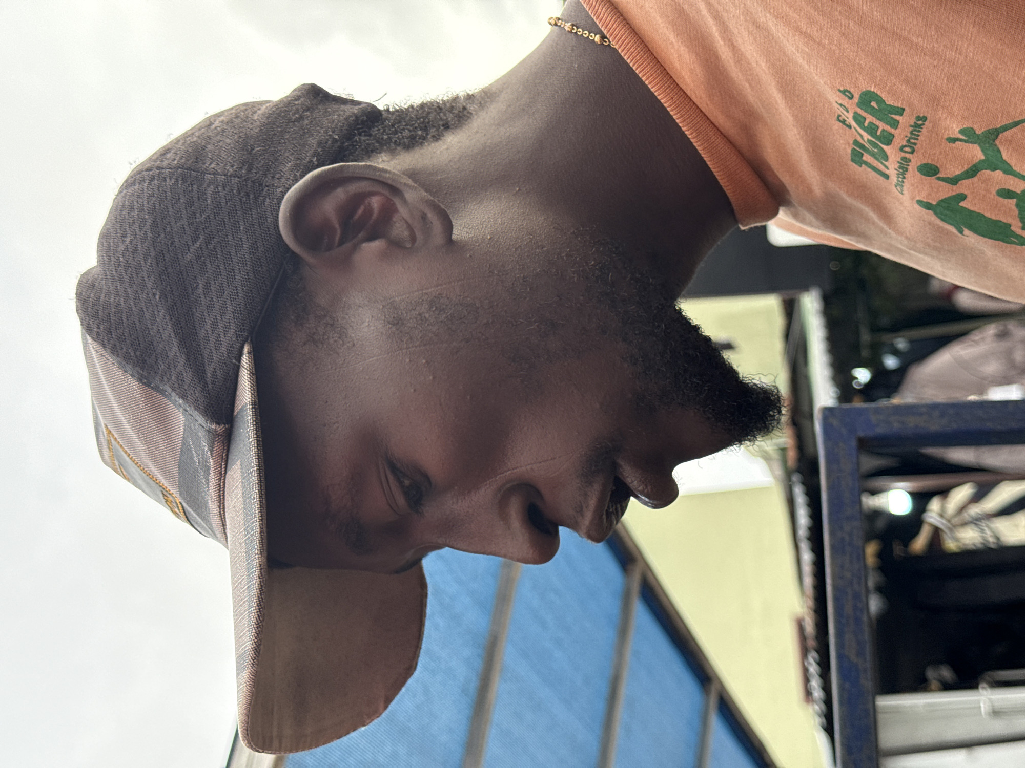 A close-up shot of a person wearing a brown cap and an orange shirt with green graphic text that includes the words "Chocolate Drinks". The person is standing outside in the city of Ado, Nigeria. A building with a blue and yellow exterior wall, and a metal structure with storage are visible in the background.