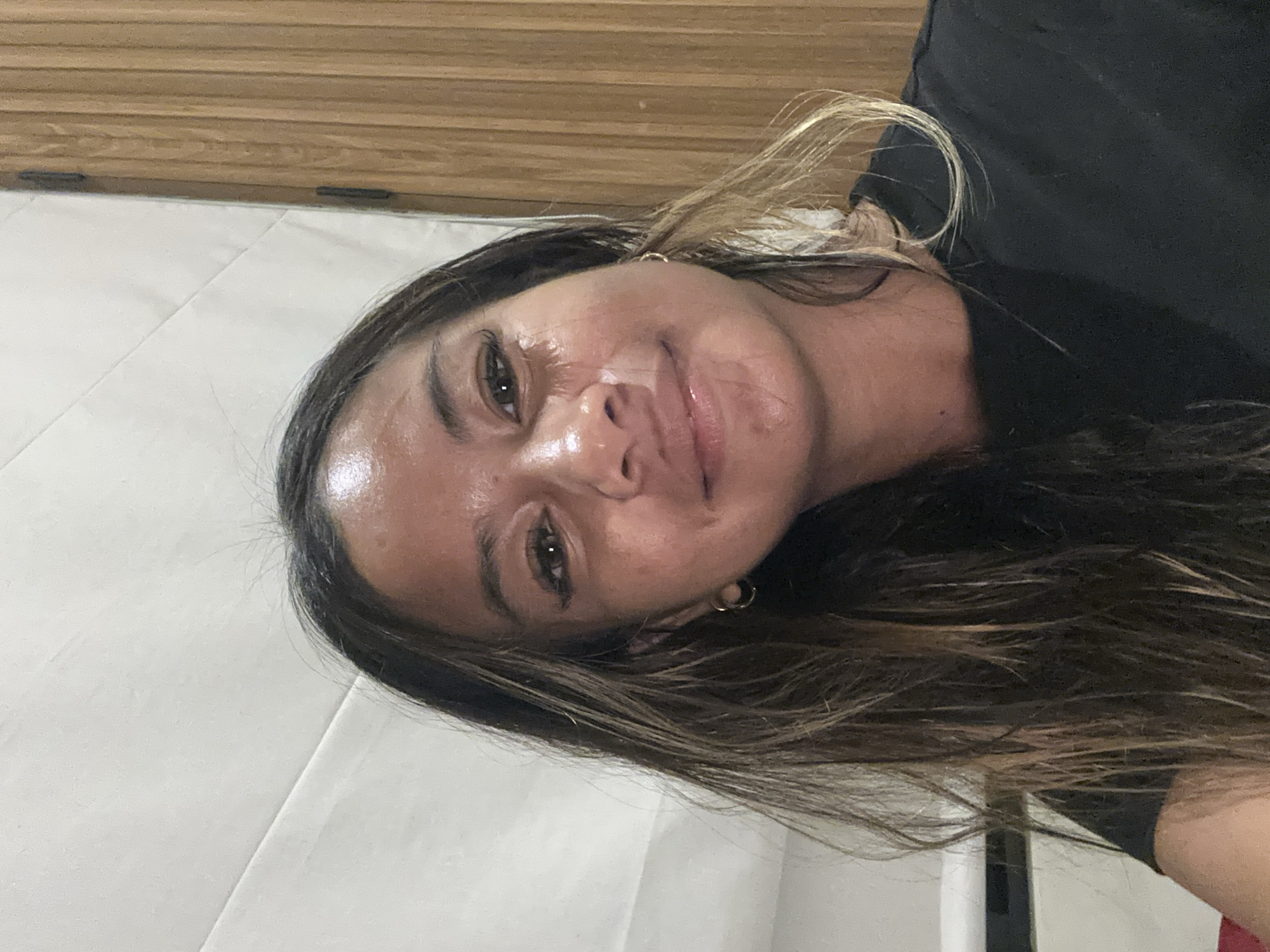 A woman is seen indoors, likely in a building in Tumbak Bayuh, Indonesia, although specific landmarks are not identifiable in the image. She is facing the camera with her head slightly tilted. She has dark hair with lighter highlights and is wearing a black shirt. The background includes a white wall and a wooden-patterned surface. The lighting is relatively bright.
