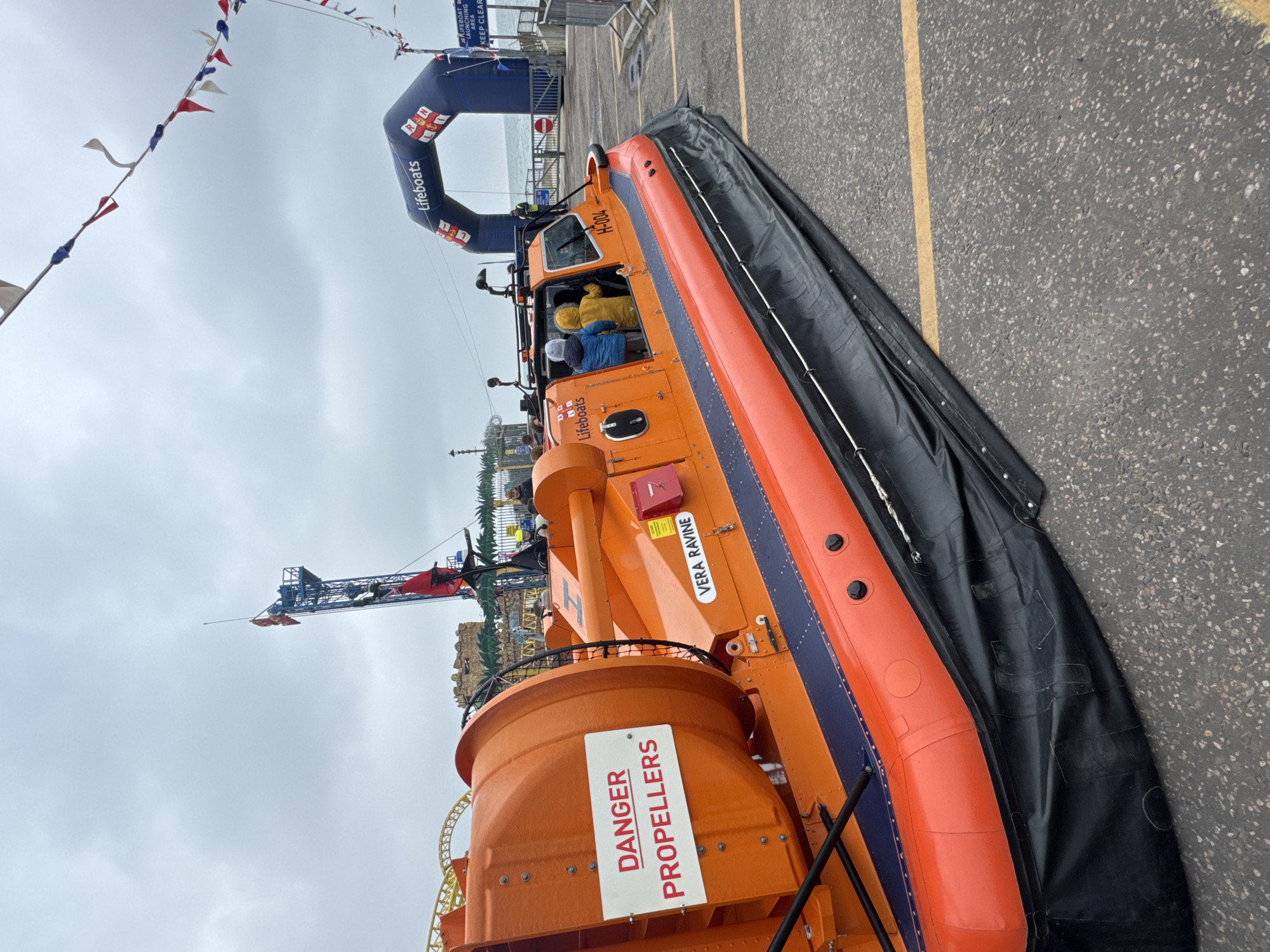 The image depicts a lifeboat hovercraft, named "Vera Ravine," parked on paved ground next to a yellow painted line. It’s likely the lifeboat belongs to the Royal National Lifeboat Institution (RNLI), as indicated by the "Lifeboats" logo on the craft and the RNLI arch visible in the background. The hovercraft is orange and blue, with a black skirt around the bottom, and features a prominent "DANGER PROPELLERS" sign. The hull is marked with "H-004." People, partially obscured, are inside the hovercraft. Pennant flags are suspended overhead. A roller coaster or amusement park ride is visible behind the hovercraft. The image was taken at a RNLI lifeboat launch area near an amusement park in Southend-on-Sea, United Kingdom. The sky is overcast.