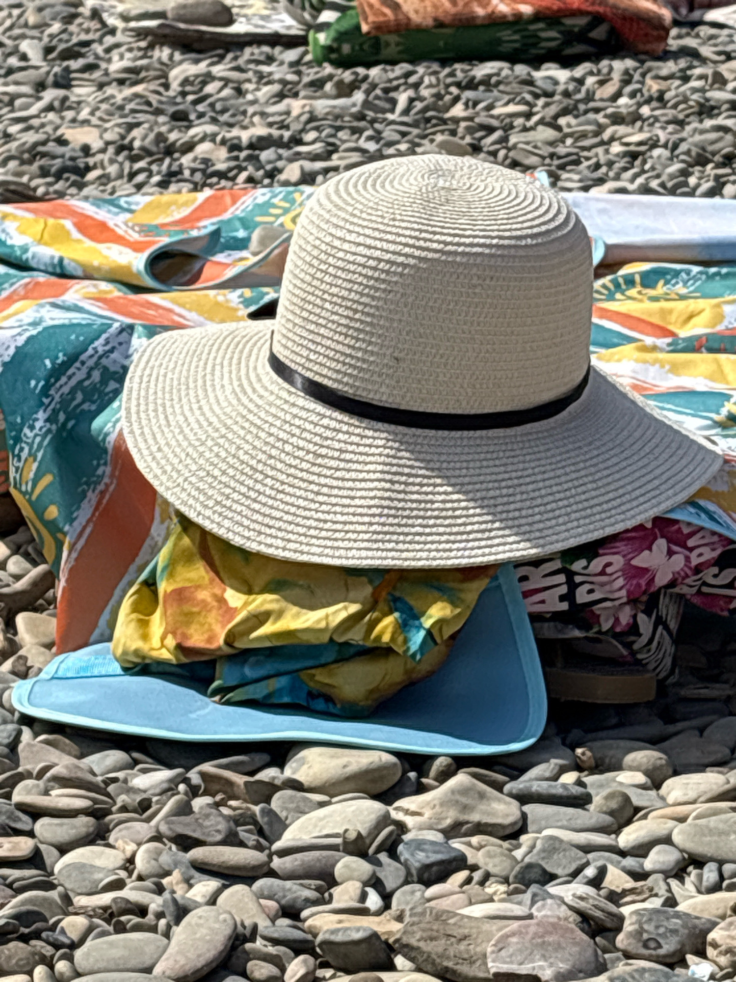 A wide-brimmed straw hat with a black band rests on top of a pile of brightly colored beach towels and clothing on a bed of grey and brown pebbles. The towels feature abstract patterns in teal, orange, yellow, and pink. A blue mat or bag is partially visible beneath the pile of items. The scene is outdoors, on a beach, with the pebbles extending into the background. The lighting suggests it is daytime.