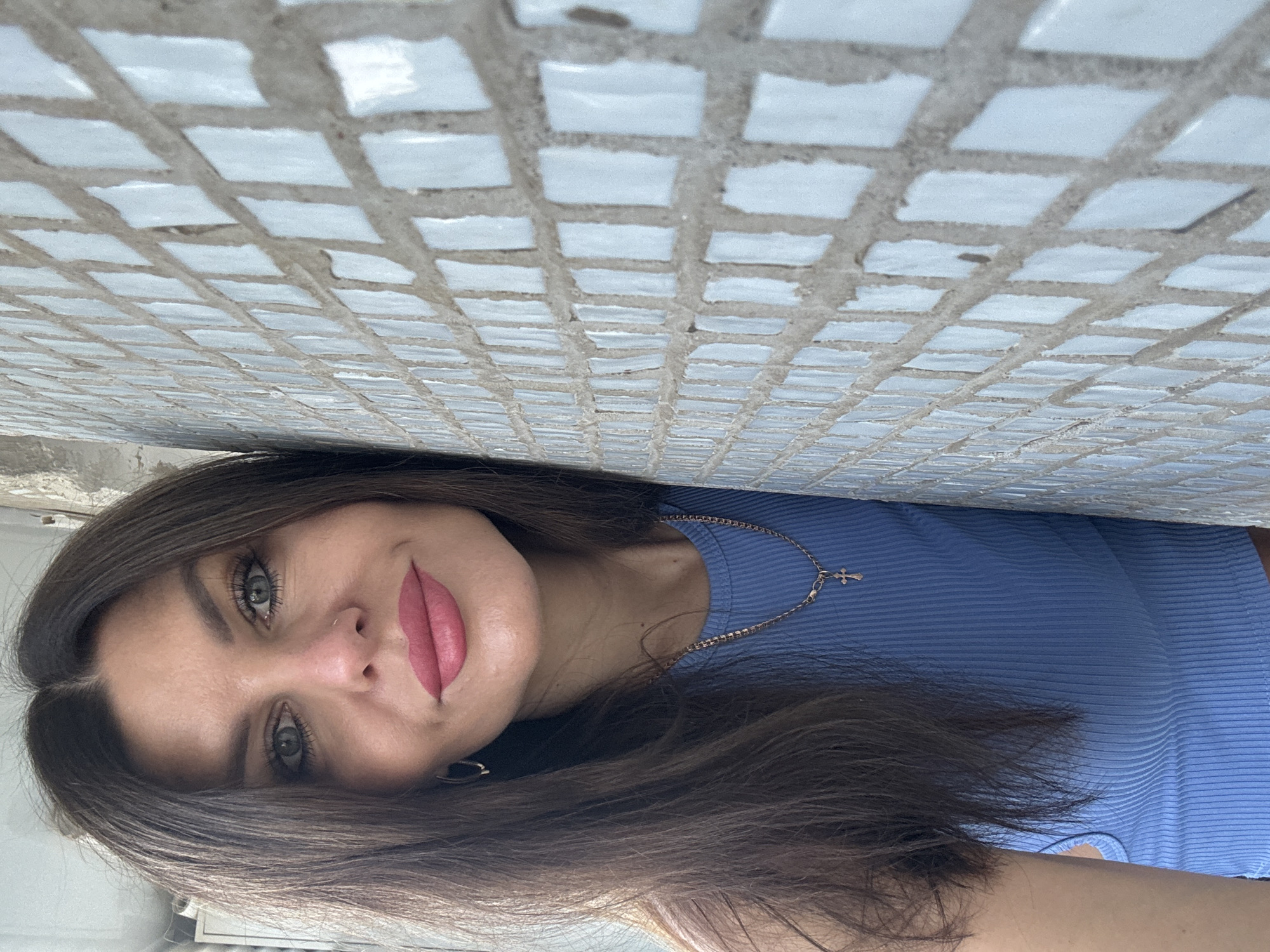 A woman with dark brown hair and full lips leans against a tiled wall. She is wearing a blue ribbed tank top and a gold chain necklace with a cross pendant. The wall is covered in small, light blue square tiles with gray grout. The background is out of focus and appears to be an outdoor setting. The image is a portrait taken from the chest up. The location is identified as Chaikovsky, Russia.