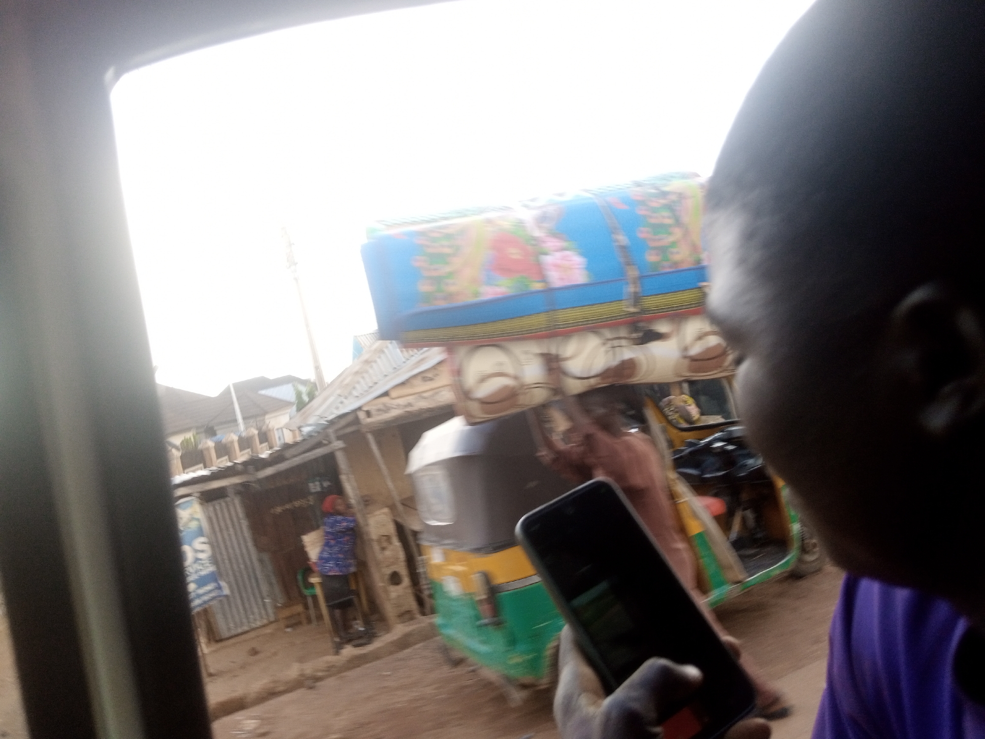 A close-up, over-the-shoulder shot captures a person holding a smartphone in the foreground, their head and shoulder dominating the right side of the frame. The phone is pointed forward, suggesting the person is taking a picture or recording a video. In the middle ground, a colorful, decorated auto-rickshaw is parked on a dirt road, loaded with goods covered by a blue and pink tarp. A woman in a blue and white patterned top and dark skirt stands near the auto-rickshaw, her back to the camera. Behind her are various market stalls with corrugated metal roofing. The background is an overexposed sky, indicating a bright day.
