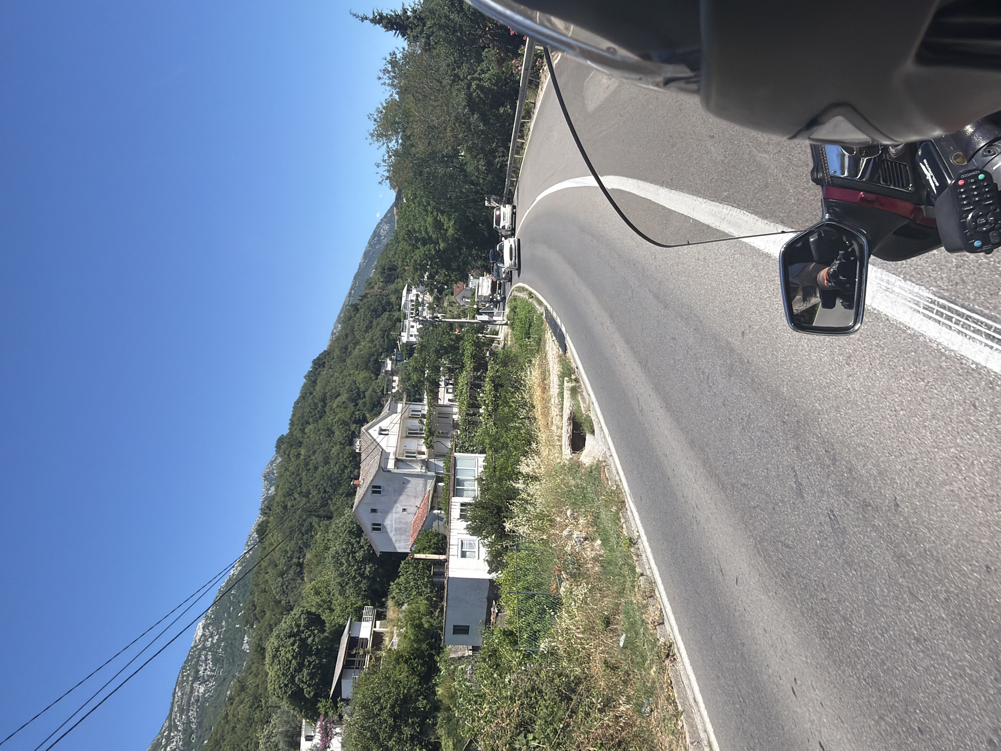 The image captures a view from a motorcycle traveling on a paved road in Mišići, Montenegro. The road curves to the right, with a white line marking its center and edge. On the left side of the road, there is a steep, tree-covered hillside with several white houses nestled among the foliage. The houses appear to be residential, with some featuring balconies and red-tiled roofs. On the right side of the road, there is a metal guardrail and more trees. In the distance, mountains rise, their peaks visible against a clear blue sky. Several cars are visible on the road ahead, including a white sedan and a white van. The foreground shows part of the motorcycle, including a windshield, a rearview mirror reflecting the rider's gloved hand and arm, and a control panel. The environment appears to be a sunny day in a rural or semi-rural area with a Mediterranean climate, given the lush greenery and the style of the houses.