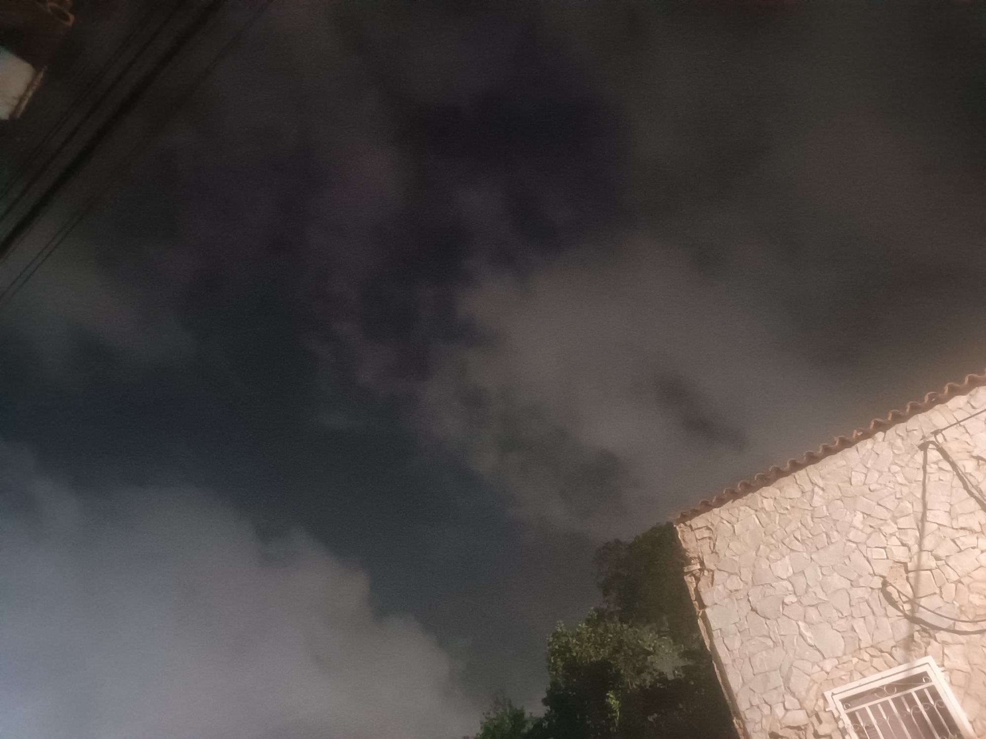 This image depicts an outdoor nocturnal scene in Porlamar, Venezuela, characterized by a dark, heavily clouded sky occupying the upper two-thirds of the frame. The clouds present a varied texture, ranging from deep grey to lighter grey, indicating atmospheric moisture. In the upper-left quadrant, multiple parallel utility lines, likely electrical or communication cables, are visible against the dark sky.
On the right side of the image, the upper portion of a building is visible. Its exterior features a light-colored, possibly beige or off-white, facade constructed from irregularly shaped, textured stone or a stone-like material. The roofline of this building, visible at the top edge of its structure, is covered with reddish-brown, semi-circular terracotta tiles. On the lower right, a portion of a white-framed window, secured with dark metal security bars, is integrated into the stone facade. Dark cables or conduits are affixed to the building's exterior on the right side.
Below the building and extending into the lower-left portion of the image, dark green foliage from trees or bushes is present. The building is illuminated by an unseen artificial light source, casting a warm glow on its facade, contrasting with the dark, unlit vegetation and sky. No individuals, specific actions, or interactions are observable in the frame.
