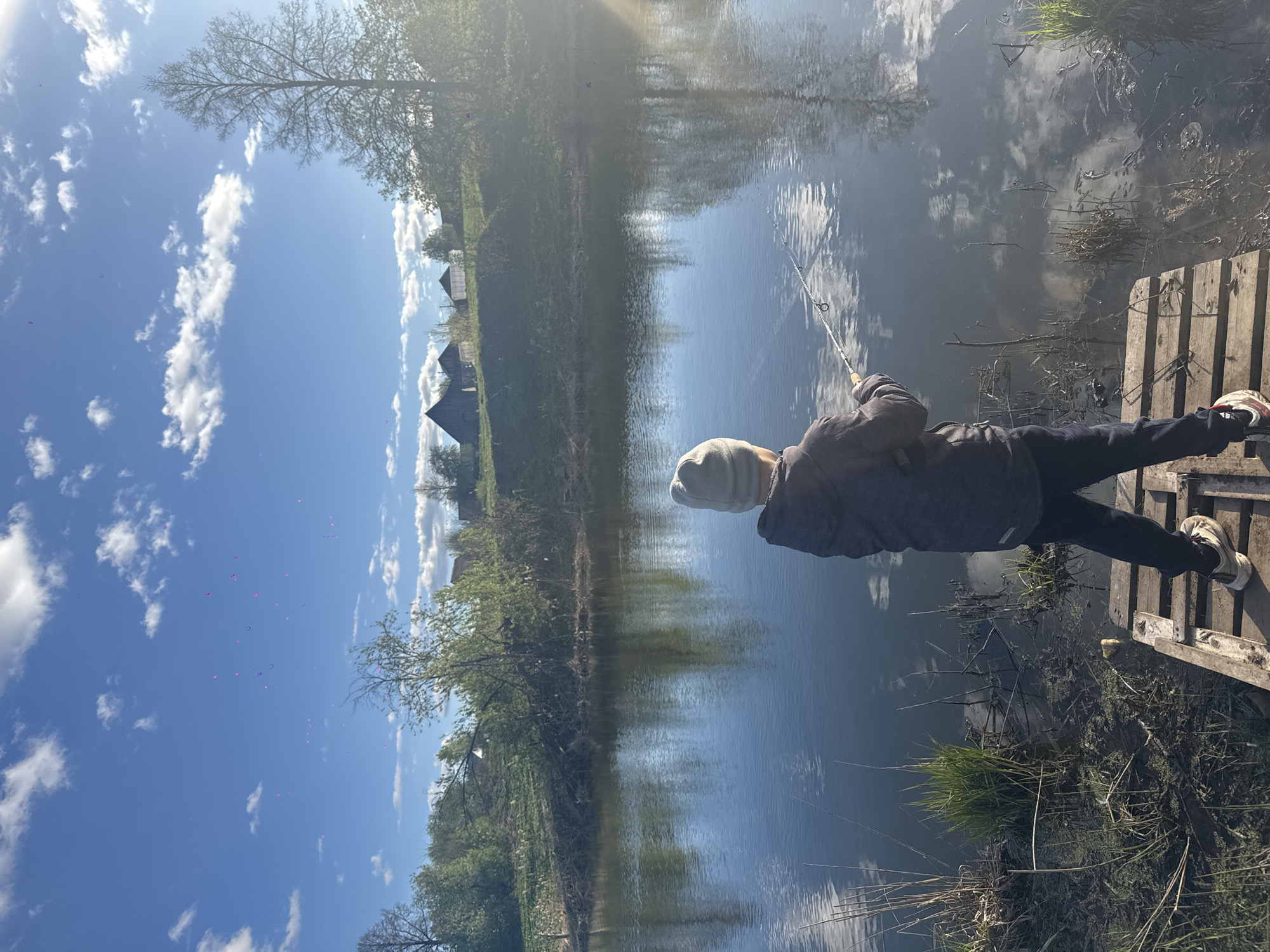 A person is fishing off a wooden dock on a body of water that likely has a significant amount of algae/vegetation, and which reflects the sky. There is a treeline on the opposite shore. The photo was taken in Ol'khovochka, Russia.