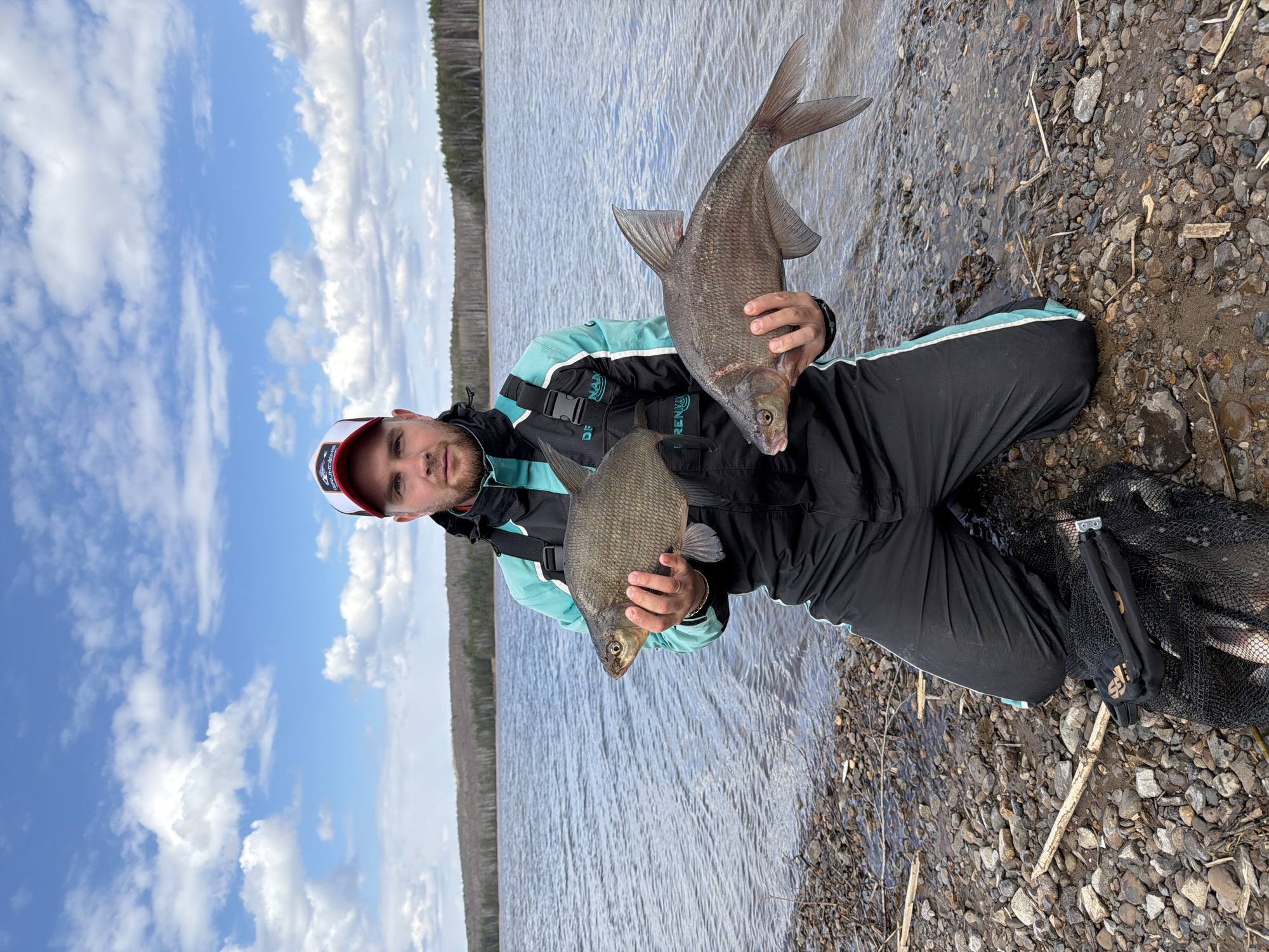 A man kneels on a pebbled shore next to a body of water with a wooded shoreline in the background. He holds two fish in his hands, one in each hand. He is wearing a black and turquoise jacket and a white and red baseball cap. A fishing net is visible near his knees. The sky is blue with scattered clouds. The location is the city of Chaikovsky, Russia.