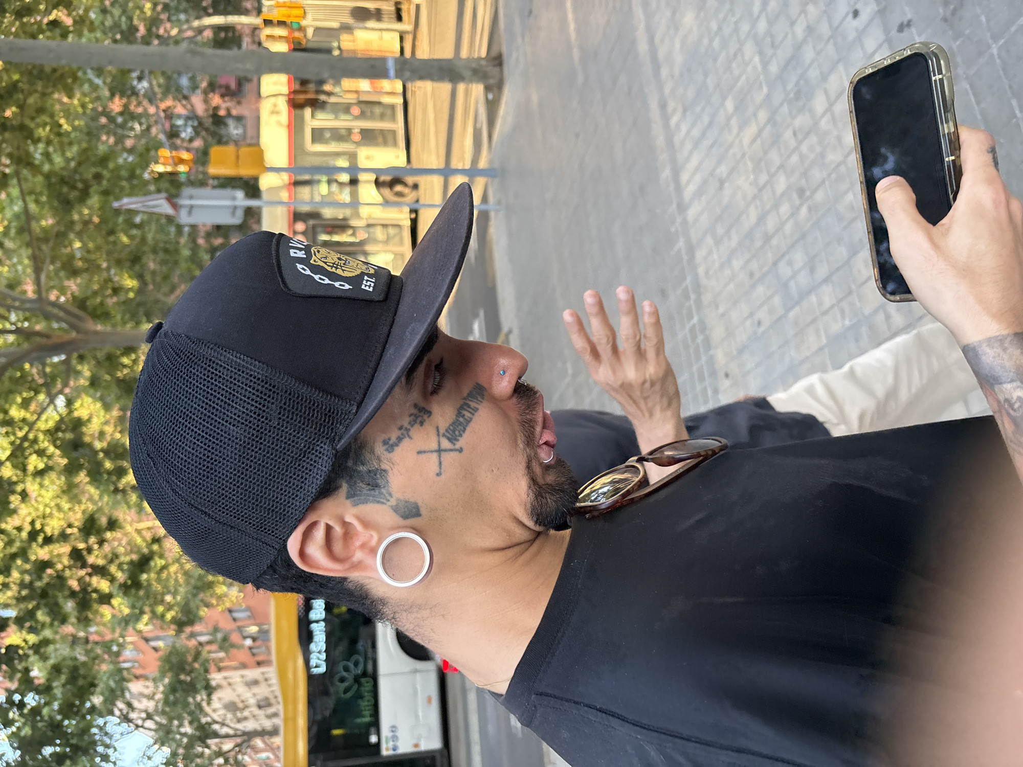 A close-up, slightly tilted image shows a person outdoors in an urban setting during what appears to be a sunny day. The subject, likely male, is seen from the chest up, facing right and away from the camera, with a black baseball cap covering part of his head. The cap features a black mesh back and a patch on the front with partially visible text "RVCA" and "EST.". He wears a black t-shirt, and sunglasses with brown frames are resting on his collar.
His face is adorned with several tattoos, including an "X" mark and script on his right cheek, and other designs around his temple area. A small stud nose piercing is visible on his right nostril, and a silver-colored ear gauge/tunnel is in his left earlobe. He has a beard/goatee. His mouth is slightly open, and his left hand is raised in a gesture, possibly waving or talking. His right hand holds a smartphone in a clear or camouflaged case, with his thumb on the screen.
In the background, out of focus, are green trees and a street scene. A yellow and white bus is partially visible, bearing the text "L72 Sant Boi" and "Hybrid," consistent with a public transport line in Barcelona, Spain. Traffic lights with yellow and red signals are also discernable further in the background, suggesting a busy street intersection. The pavement or sidewalk beneath the subject is textured. The overall impression is a casual, candid street shot.