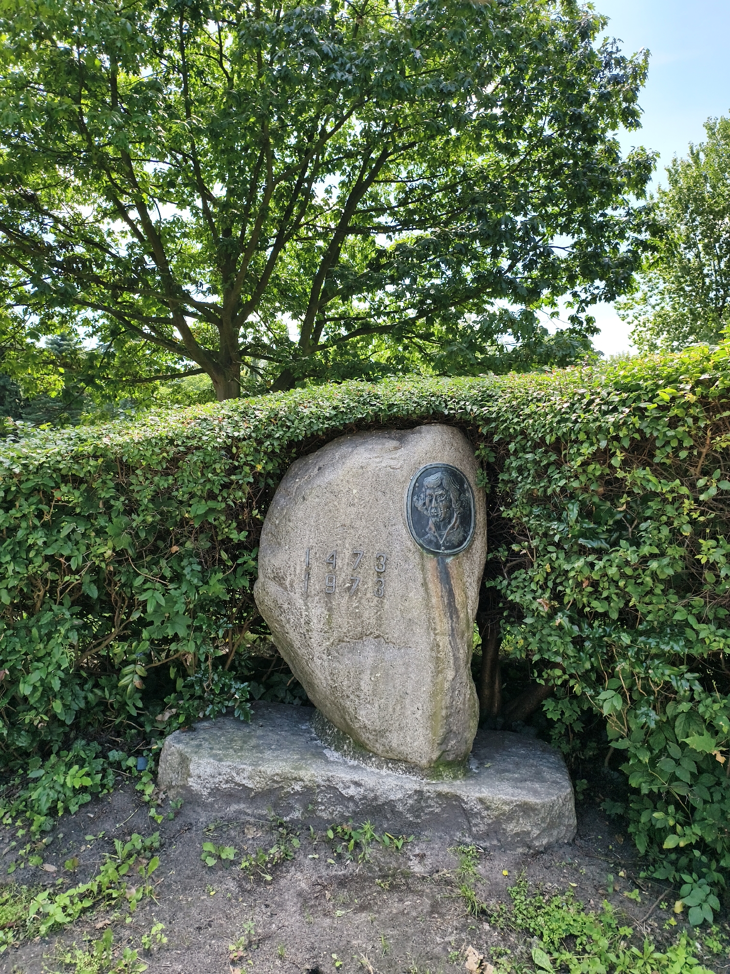 The image shows a large, grey stone monument set within a neatly trimmed green hedge. The stone has a round, bronze plaque with a profile portrait of a man attached to its upper right side. Below the portrait, etched into the stone, are the numbers "1 7 3 3". The monument is resting on a larger, flatter grey stone base, which is partially covered by grass and dirt. The background consists of a lush green tree with visible branches and leaves, and a bright blue sky. This monument is located at the Henryk Dende Park in Tczew, Poland, commemorating the mathematician and astronomer Henryk Dende, who was born in 1733.