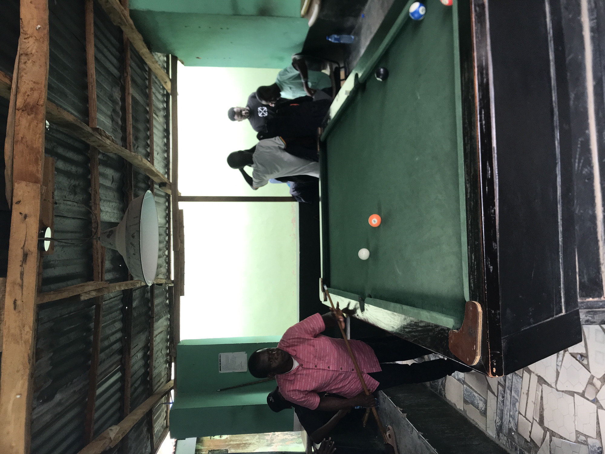 This image captures a lively scene inside a rustic pool hall, likely near New Stadium in Nigeria, during daylight hours. The main subject is a man in a pink striped shirt and dark trousers, actively playing pool. He is leaning over a green-felted pool table, holding a cue stick, intently focused on hitting the white cue ball. An orange ball, a blue ball, and a black ball are visible on the table.
Several other men are gathered around the table in the background, observing the game. One man wearing a black t-shirt with a white, possibly geometric, graphic is clearly visible. The overall atmosphere appears casual and social, typical of a community recreational spot.
The setting is characterized by its simple, utilitarian construction. The walls are primarily made of corrugated metal sheets, supported by rough wooden beams. Some interior wall sections are painted a light green. A large, industrial-style dome light fixture hangs above the table area, supplementing the natural light streaming in from a bright opening to the right of the frame. The floor near the pool table is covered with a mosaic pattern of light-colored tiles. A white rectangular sign with unreadable black text is affixed to a green wall behind the player, possibly displaying game rules or local information.