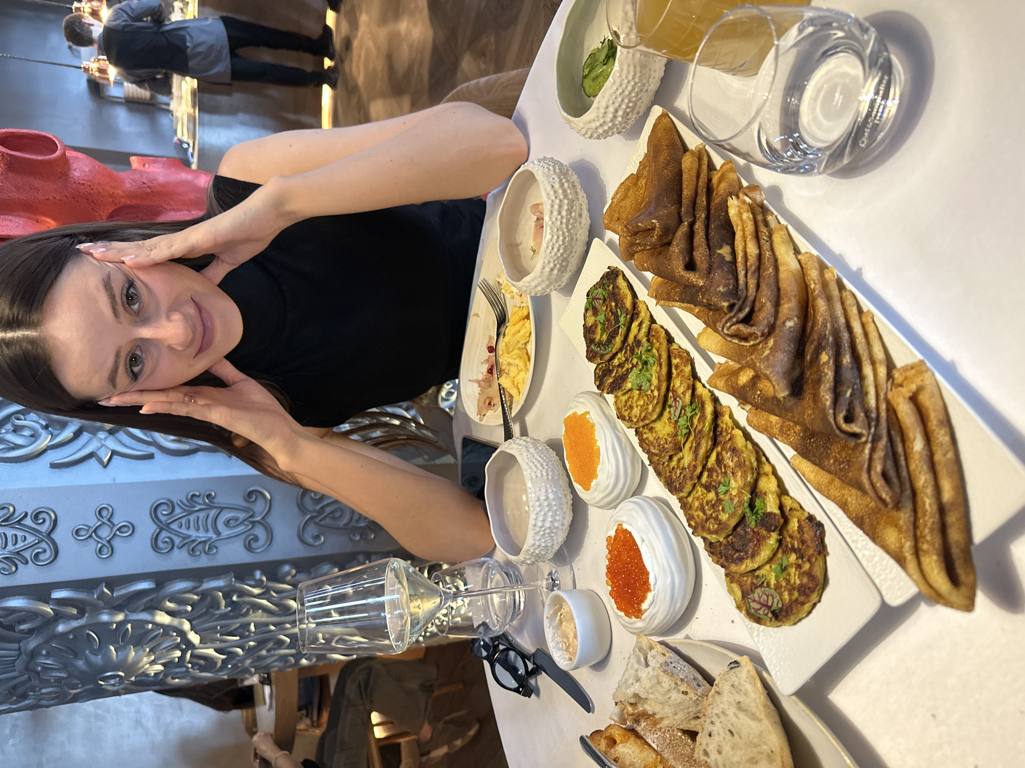 A young woman sits at a table filled with food in what appears to be a restaurant in Krasnoyarsk, Russia. The table is covered with various dishes, including pancakes, fried zucchini, caviar, bread, and drinks. The restaurant's interior features ornate decorations and a silver wall panel. Other people can be seen in the background, including a waiter. The woman is looking at the camera with her hands near her face. A pair of eyeglasses are on the table near her.