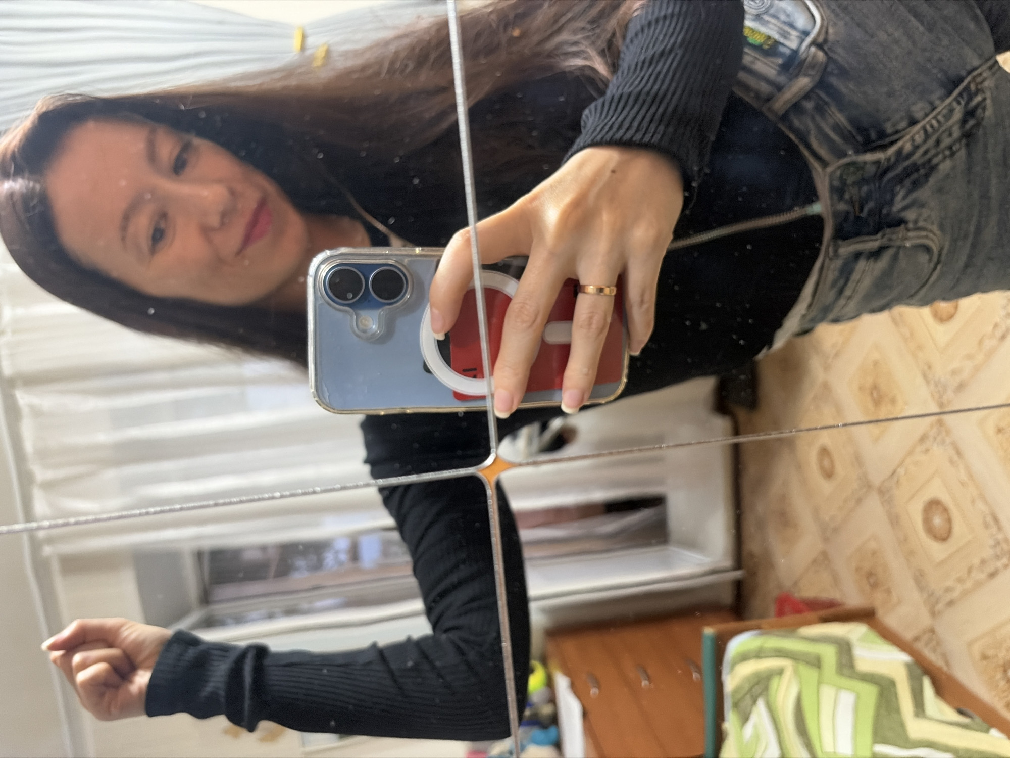 A woman is taking a selfie in a mirror. She is wearing a black long-sleeved shirt and blue jeans. She is holding a smartphone in her right hand. Her left arm is raised in the air. The background shows a bedroom with a bed and a dresser. The floor is tiled.