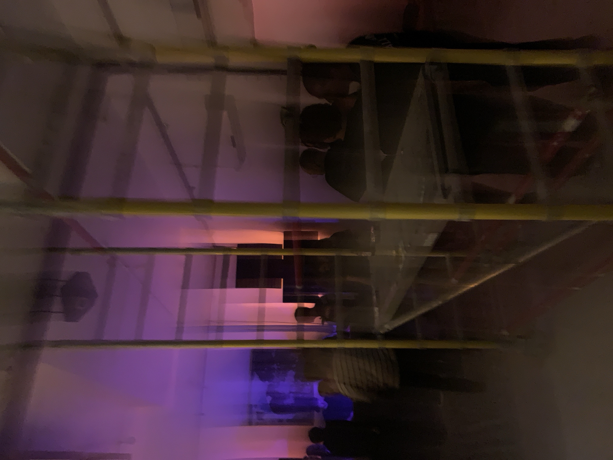 The image shows an interior space in Tel-Aviv, Israel, viewed through a metal scaffold. The scene appears to be a dimly lit room with purple and orange ambient lighting. Several people are present in the background, some walking or standing. The scaffold has yellow horizontal supports. A speaker is attached to the wall in the background. The image has motion blur.
