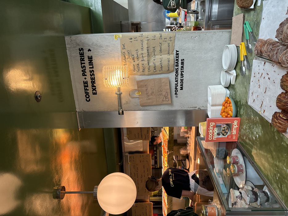 The image displays the interior of a food service establishment, identified as The Corner Shop by Populations, located at 125 Whitechapel Road, London, United Kingdom.
The scene focuses on a counter and an adjacent preparation area. In the foreground, a green marble-patterned counter presents an array of pastries, including croissants, arranged on metal trays. A red cookbook titled "cooking made simple" stands upright. Adjacent to it are a white bowl filled with small orange fruits (likely kumquats), stacks of white plates, and white disposable containers. Tongs with yellow and green handles rest on the counter surface. Behind a clear glass barrier, several small jars and individual dessert portions in clear plastic containers are displayed.
To the right, a prominent concrete or metallic pillar features black sans-serif text indicating "COFFEE + PASTRIES EXPRESS LINE" with a directional arrow pointing right. Below this text, a decorative light fixture with a ribbed, translucent shade emits a warm glow. Two brown paper bags are pinned to the pillar; one advertises "GRAPEFRUIT JUICE £6," and the other displays a "BEVERAGE BAR JUICES" menu with listed prices for various fresh juices, including "Fresh Apple," "Fresh Orange," "Fresh Blood Orange," "Organic Celery," "Carrot + Ginger," "Turmeric + Orange Shot," and "Homemade Lemonade." The pillar also displays "@POPULATIONS.BAKERY MADE UPSTAIRS."
In the mid-ground, a female staff member, wearing a dark t-shirt with "CORNER SHOP" printed on the back and a white apron, stands with her back to the camera behind a service counter, appearing to be working. Further in the background, a second female staff member is partially visible, also engaged in tasks within a kitchen or preparation area. Trays of prepared food, potentially flatbreads or pizzas, are visible under warm lighting in this background section. The ceiling is a reflective, dark green surface, from which a large, spherical white pendant light fixture hangs on the left side of the frame. Additional handwritten menu boards are suspended in the distant background.