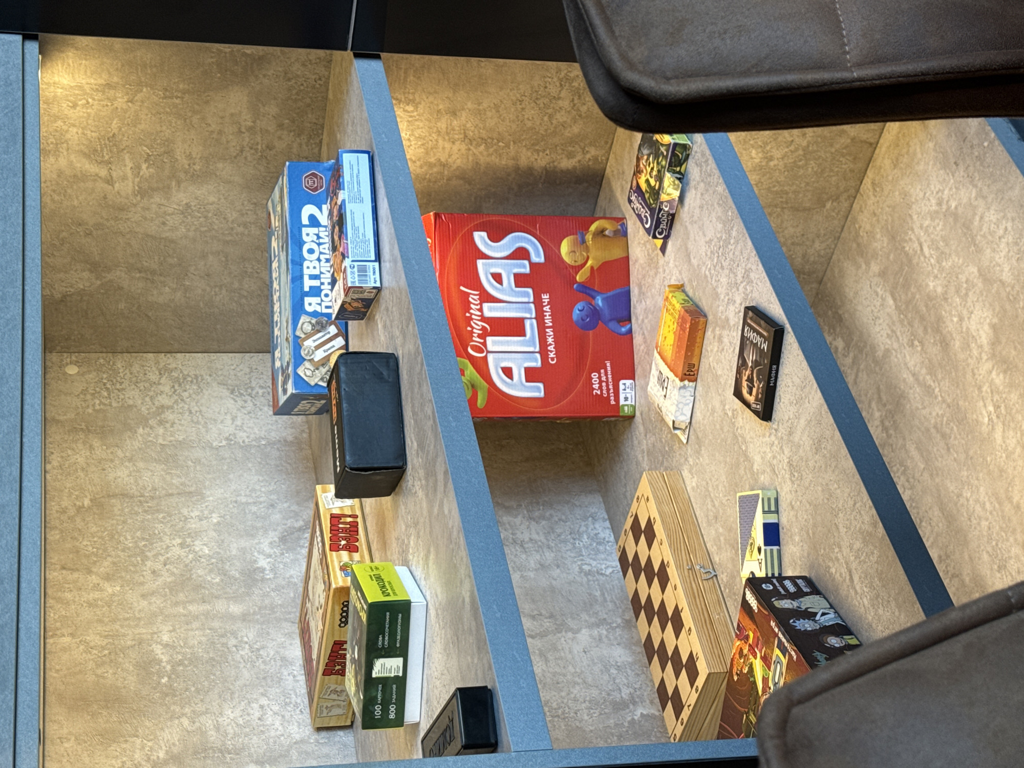 A high-angle, close-up shot shows the interior of a shelf, likely within a cabinet or storage unit. The shelf is divided by vertical blue dividers. On the left side is the board game "Crocodile", "I Understand You 2" and "Banzai" the board game, some small rectangular black boxes. On the right side is "Alias", a chess set, a card with blue and yellow design and some small board games "Mafia" and "Svoya igra". There is a chair in the foreground, with parts of the seat cushion visible on the right and top edges. The setting seems to be indoors, in a home or office environment. The presence of Russian board games suggests the location may be Russia. The brands of the board games also suggest the location is Perm, Russia.