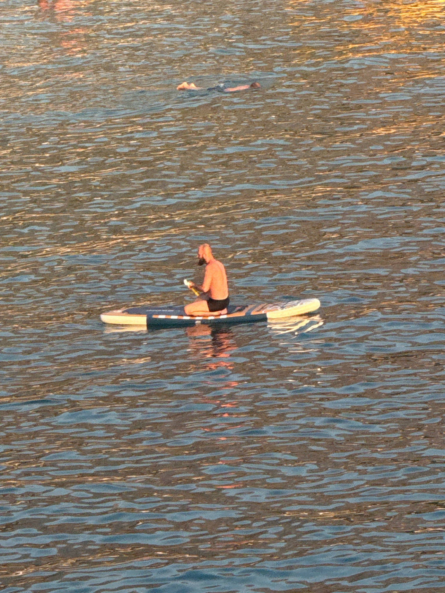 A man is paddleboarding on the water in Kamenovo, Montenegro. He is kneeling on a paddleboard and holding a yellow object in his hands, which appears to be a phone or a book. The water is choppy and reflects the sunlight, creating a shimmering effect. In the background, another person is swimming. The sky is not visible in the image.