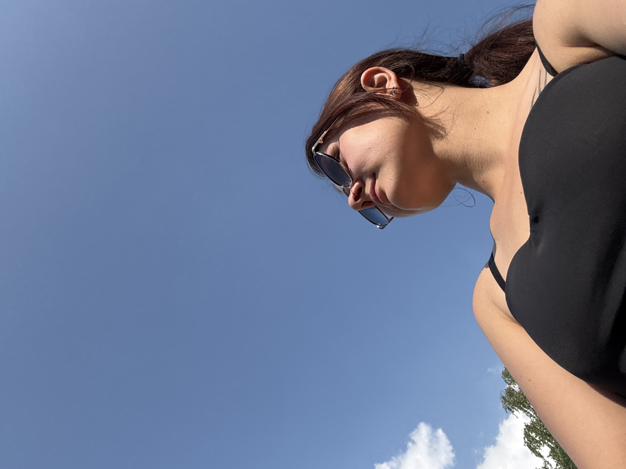 A young woman wearing sunglasses and a black tank top is pictured from a low angle against a bright blue sky with a few clouds. She has her hair pulled back in a ponytail and is looking upwards. The scene appears to be outdoors, likely during the daytime. The location is not specified within the image or context.