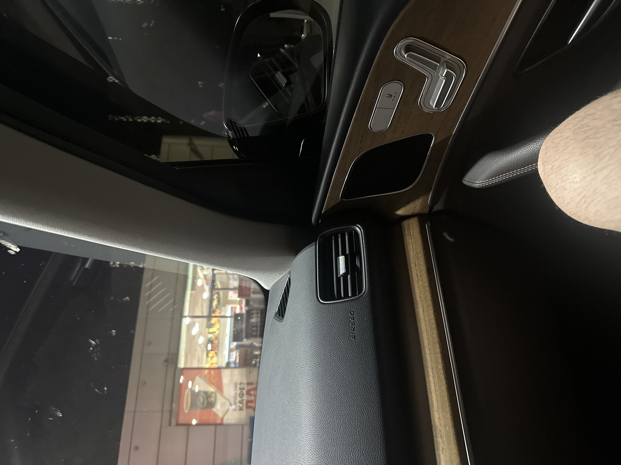 The image shows the interior of a car, looking out through the passenger side window. Outside, a brightly lit building with large windows is visible, featuring advertisements and shelves stocked with products. A sign with Cyrillic text, including "КАФЕ?", is prominent. The car's dashboard, air vent, and a portion of the center console with wooden trim and seat adjustment controls are visible inside the car. A person's leg is also in the frame. The scene suggests the car is parked outside a store or cafe in Ruse, Bulgaria, likely at night due to the lighting.
